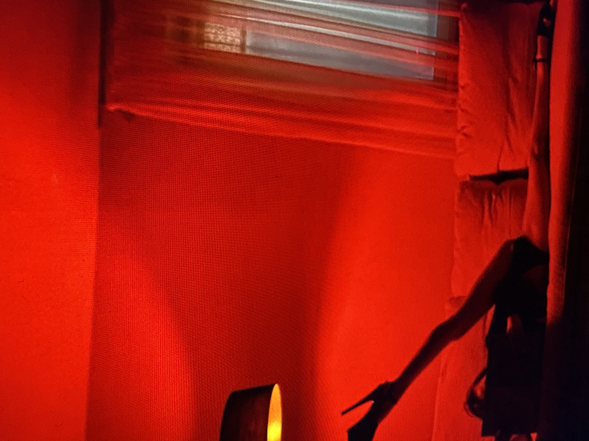 A person, rendered in silhouette, lies on their back on a red couch. They are wearing a black garment and black high-heeled shoes. One leg is extended upwards at a sharp angle, while the other is bent. The scene is illuminated by a strong red light, casting a warm glow over the room. A black lampshade is visible to the left, and sheer curtains cover a window on the right. The overall impression is one of intimacy and drama, with the red lighting creating a striking visual effect. The location is Unknown, Unknown.