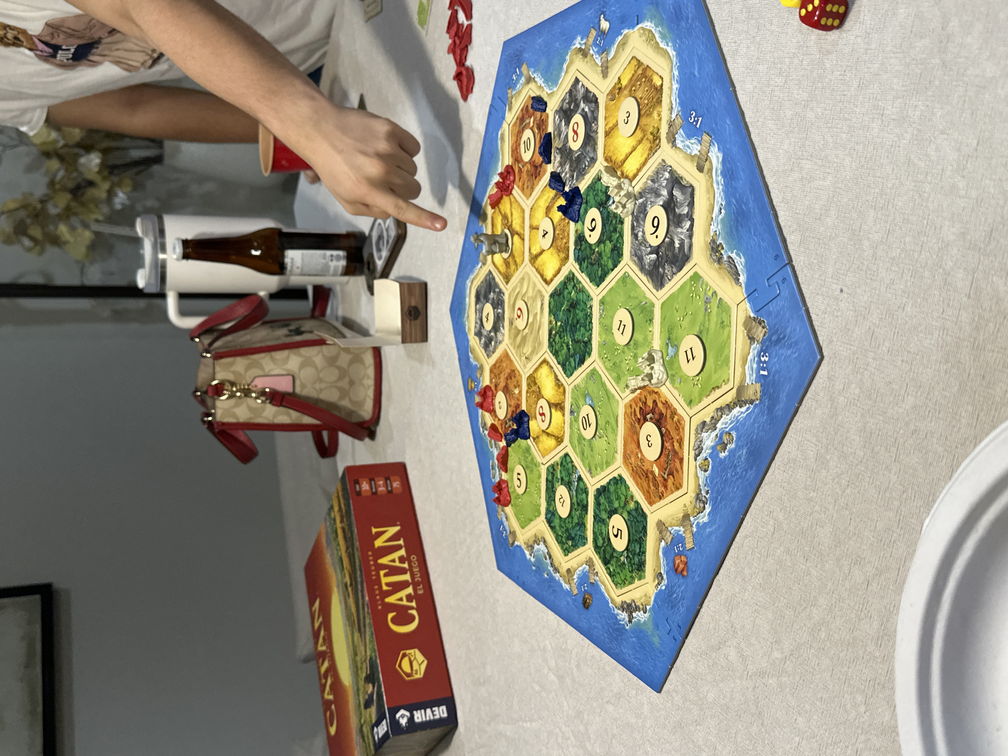The image shows a tabletop game of Catan being played on a light-colored tablecloth. The game board, composed of hexagonal tiles depicting various terrains, is prominently displayed. Several pieces, including red and blue settlements and roads, are placed on the board. A person's hand is visible, with a finger pointing towards a piece on the board, indicating an action or decision in the game. To the left of the board, a box of the Catan game is visible, along with a handbag. In the background, there's a white insulated mug, a glass bottle, and a person wearing a white t-shirt. A red cup and a yellow and red die are also present on the table. The lighting suggests an indoor setting, possibly a home or a casual gathering place. The specific location in San Bernardino Tlaxcalancingo, Mexico, cannot be determined from the visual information provided in the image.