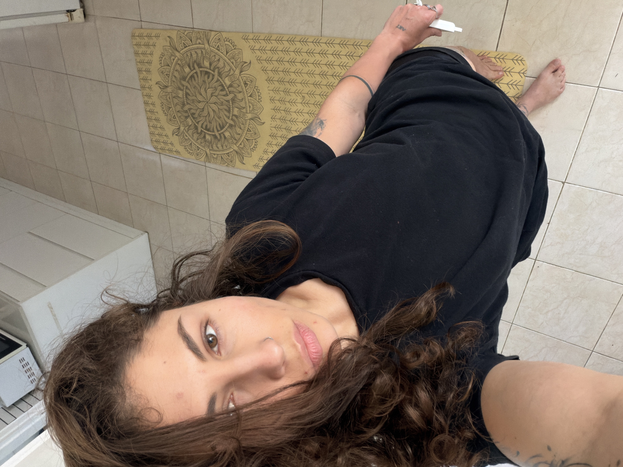 An angled, close-up selfie shows a woman indoors in Kiryat Bialik, Israel, looking directly at the camera. She has long, dark brown, curly hair that frames her face, brown eyes, and a neutral expression with a subtle nose piercing. She is wearing a black top or dress.
Her right wrist features a visible "ART" tattoo, while her left arm displays a solid dark band tattoo and another design near her elbow. Her bare feet are visible, resting on a light-colored tiled floor and partially on a yellowish-tan yoga mat. The mat is adorned with an intricate circular mandala design on one end and repeating arrow-like patterns across its length. In her right hand, she holds a small white tube-like object.
In the background, partial views of white household appliances, including what appears to be a microwave oven, are visible against the tiled wall, suggesting an indoor setting like a kitchen or living area.