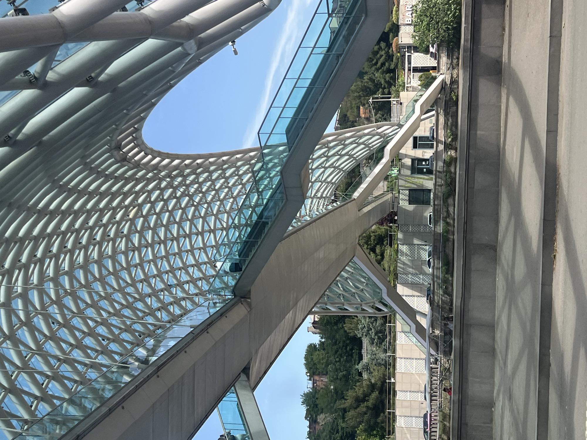 The image shows the Bridge of Peace in Tbilisi, Georgia. The bridge has a modern design, featuring a steel and glass structure with a curved, web-like canopy. There is a pedestrian walkway extending across the bridge, lined with glass railings. A street and buildings can be seen alongside the bridge. The background includes trees and vegetation, typical of an urban environment. The sky is visible between the steel structure, with clear blue color.