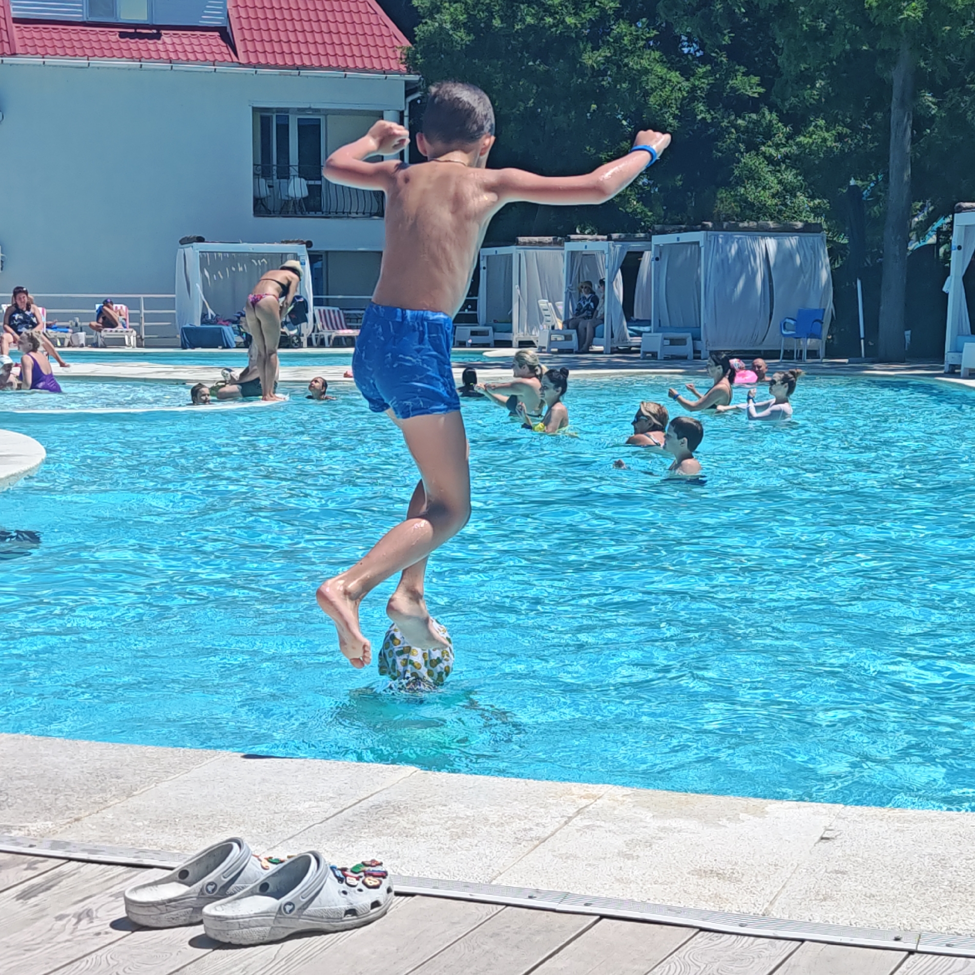 In this media file, a young boy is captured mid-leap in a bright blue swimming pool. He is wearing blue patterned swim shorts and has a blue band on his left wrist. The boy appears to be jumping from something on his right, possibly a diving board or a inflatable. His arms are outstretched and his fists are clenched, suggesting a moment of exertion or excitement. His body is wet, indicating he has been in or around the water.
The pool is surrounded by a white tiled edge and a wooden deck. In the foreground on the deck, a pair of light gray Crocs are placed side-by-side.
In the background, other people are visible in the pool, some swimming and others standing. Behind the pool, there is a white building with a red tiled roof and several white cabanas with draped curtains. Lush green trees are visible behind the building, suggesting an outdoor setting on a sunny day. The sunlight is bright, casting shadows and illuminating the water.
The overall scene depicts a recreational activity at a swimming pool, likely during warm weather. The boy's pose and the surrounding atmosphere suggest a lively and energetic environment. There is no visible text in the image.