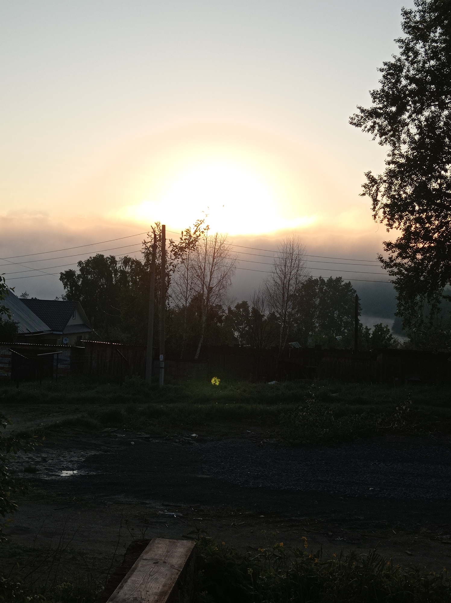 The photograph depicts an outdoor scene in Degtyarsk, Russia, at what appears to be either sunrise or sunset. The sun is positioned high in the frame, obscured by clouds, creating a diffused glow. The silhouette of a house and fence line the left side of the frame. Utility poles with power lines cross the upper portion of the image. Trees are visible behind the house and fence, with some obscured by what appears to be fog or mist. The foreground shows a dimly lit dirt road, with puddles and vegetation present. A wooden plank is visible on the lower left. The lighting suggests a low-light environment, with the lower part of the image in near darkness. The overall setting is rural or suburban. No people or other actions are visible in the picture.
