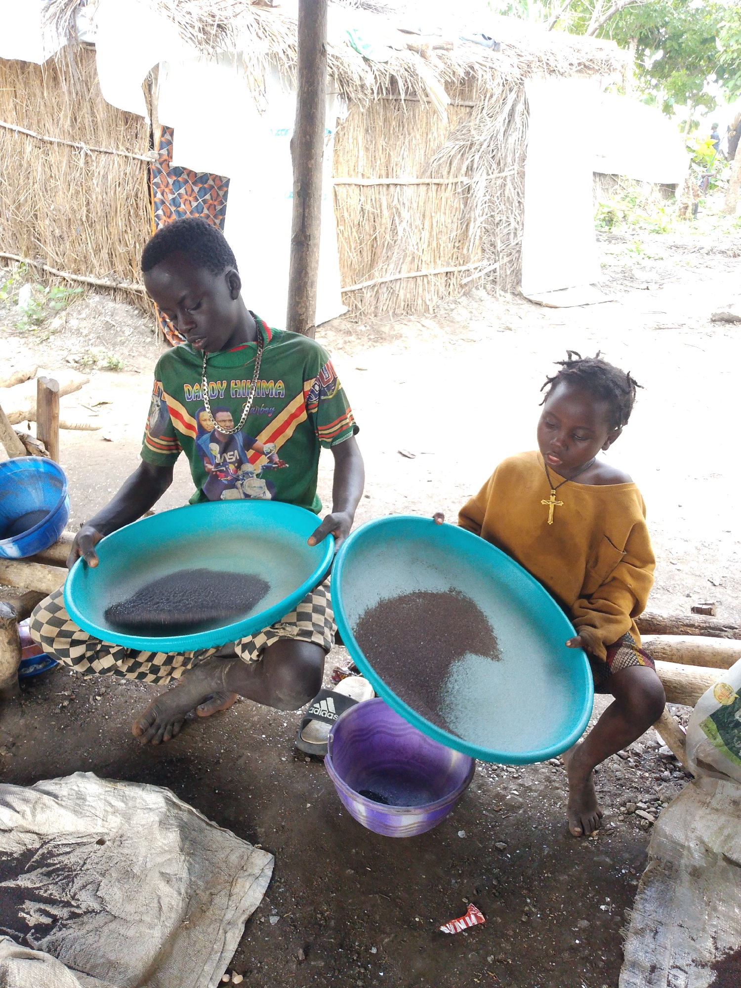 In the city of Unknown, Unknown, two young boys are engaged in what appears to be gold panning. The scene is set outdoors, with a rustic shelter made of thatch and wood in the background. One boy, wearing a green t-shirt with a gold chain and a graphic print, sits on the ground holding a large blue basin filled with dark soil. He is looking down at the basin, seemingly sifting through the contents. His checkered pants are visible. Beside him, to the right, another boy, wearing an oversized mustard-colored sweater and a small patterned skirt, holds a similar blue basin. He also has a gold cross necklace. His gaze is directed downwards, focused on the material in his basin. A purple bowl sits on the ground between them. In the foreground, a large piece of crumpled gray fabric is spread out, and various other objects, including a blue bucket and what appears to be a discarded packet, are scattered around the damp, earthy ground. The overall impression is one of laborious, possibly subsistence-level work in a rural or underdeveloped setting.