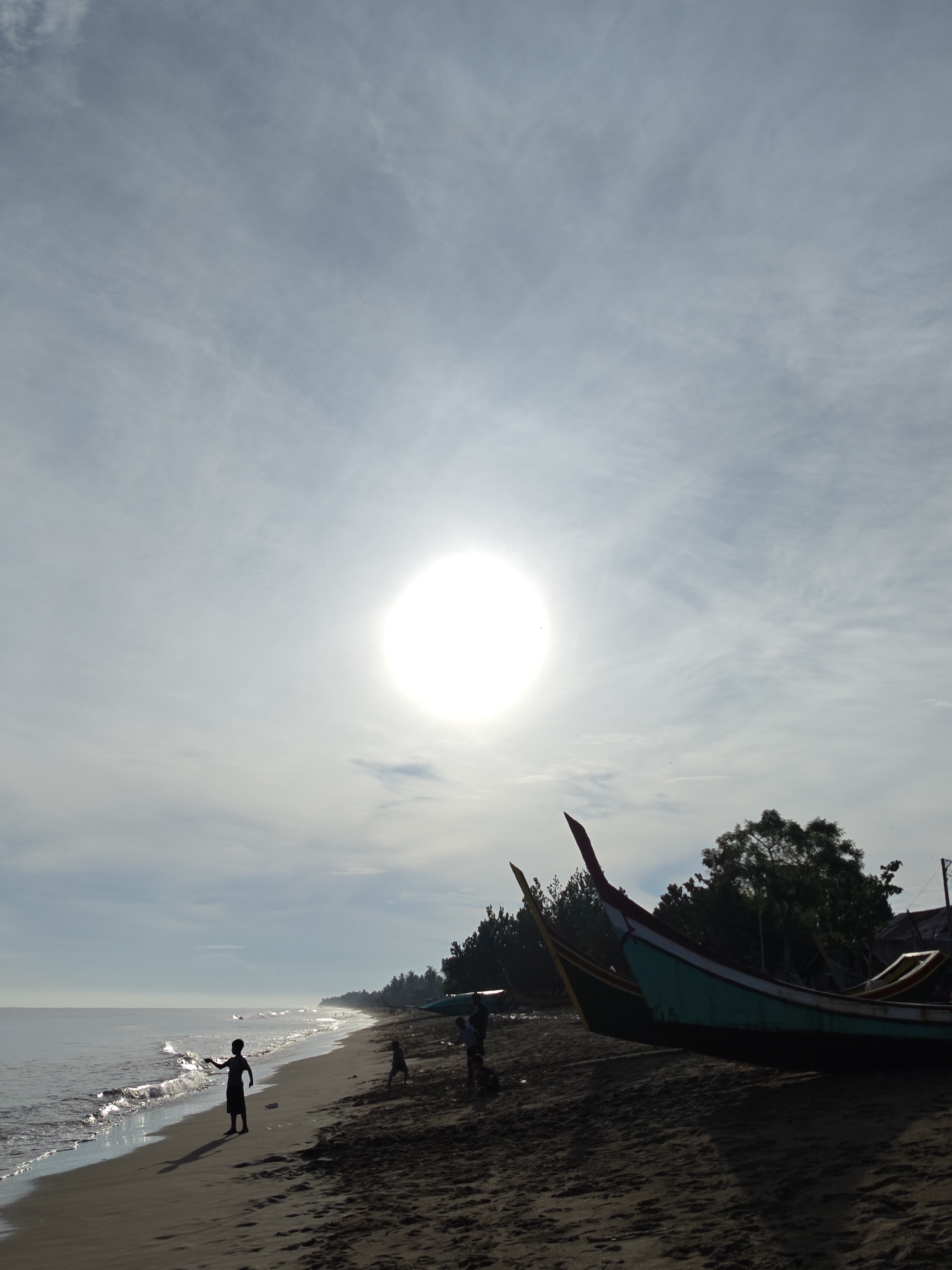 A bright, round sun is visible in a hazy sky above a sandy beach in Curehtunong, Indonesia. The sun is the brightest point in the image, partially obscuring the sky with its glare. The sea is calm with gentle waves lapping at the shore. On the beach, a lone child stands silhouetted against the water. Further down the beach, other figures, possibly children and adults, are present, though less clearly defined. Two fishing boats are pulled up onto the sand, one a vibrant teal and red, the other a duller yellow. Behind the boats, a line of trees forms a dark green backdrop against the sky. The overall impression is one of a serene, early morning or late afternoon scene.