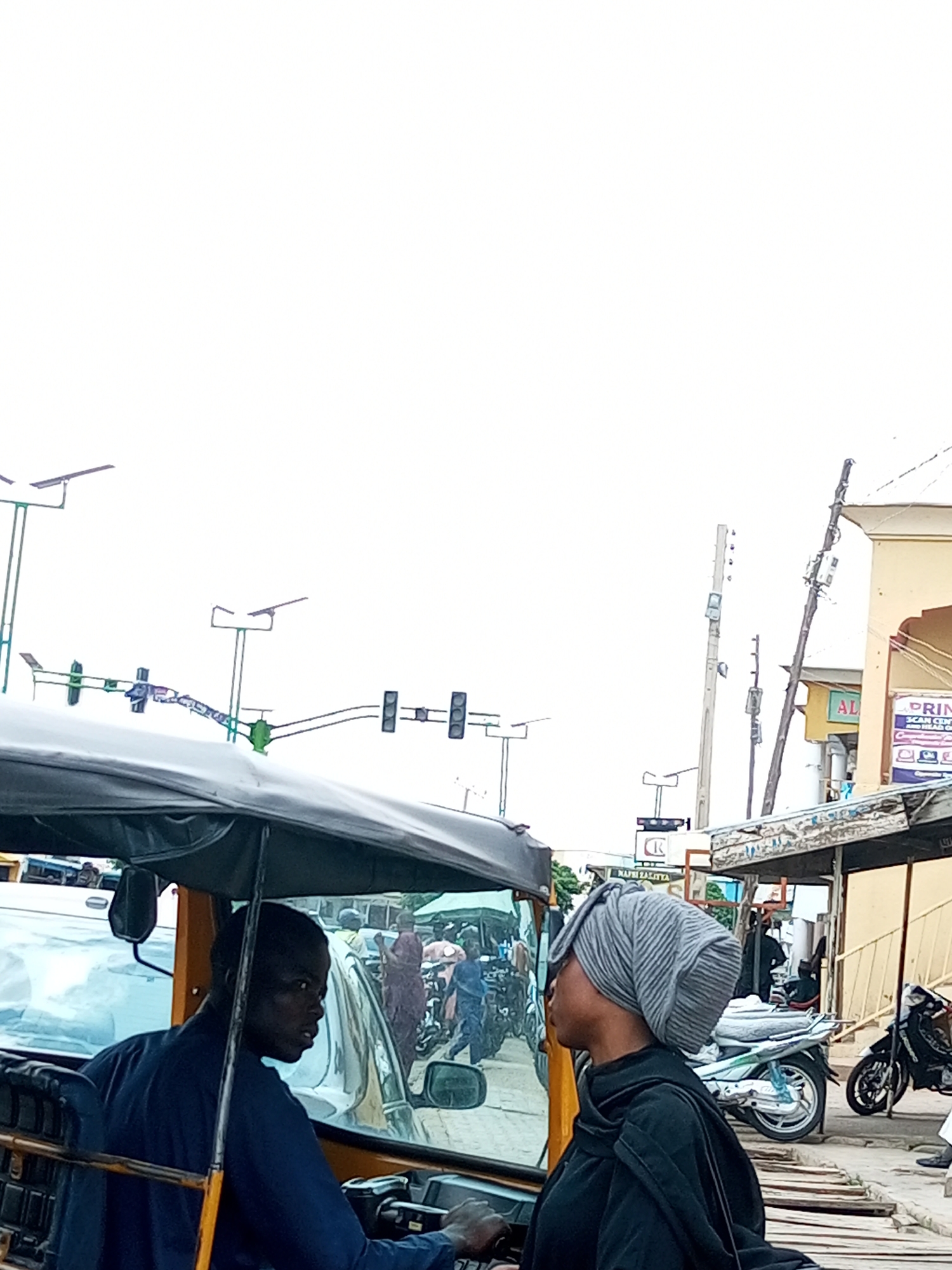 The image captures a street scene in Arawa, Nigeria, with a focus on a yellow motorized tricycle in the foreground. A male driver, dressed in a dark blue shirt, is seated in the driver's position, looking to his right. Beside him, a female passenger, wearing a grey headscarf and a black garment, is also looking to her right, away from the camera. The tricycle has a dark canopy extending over the passenger area.
In the background, traffic signals are visible, some appearing to be malfunctioning or modified with additional lights. There are also poles with lights and what appear to be telecommunication equipment mounted on them. Buildings line the street, with one structure featuring a sign that reads "AL" and another with "PRIN" and "SCAN CENTER." Several motorcycles are parked or moving in the background, along with other people and vehicles, indicating a busy urban environment. The overall atmosphere suggests a typical day in a Nigerian city, possibly at a busy intersection. The sky is overcast.