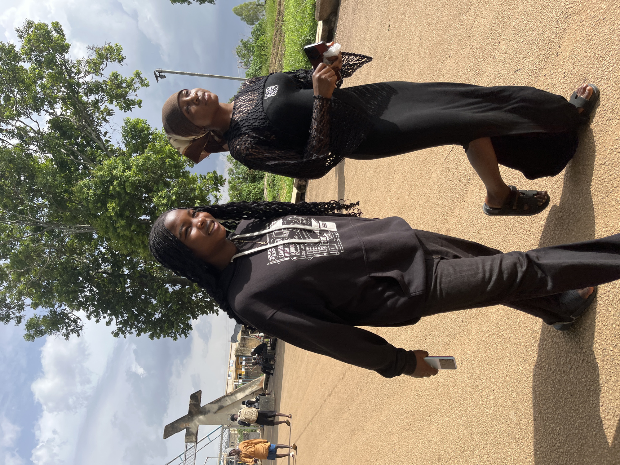 Two young women walk side-by-side in front of a large tree with a concrete cross structure behind them in the city of Jos, Nigeria. The woman on the left wears a black hoodie with a white graphic and black pants. The woman on the right wears a black dress with a lacy overlay on the sleeves and a brown headscarf. The woman on the right is holding a white object and a phone in her hands. In the background, other people are seen walking and sitting, and a street lamp is visible. The ground is a light brown, sandy surface. The sky is partly cloudy with a mix of sun and shade.