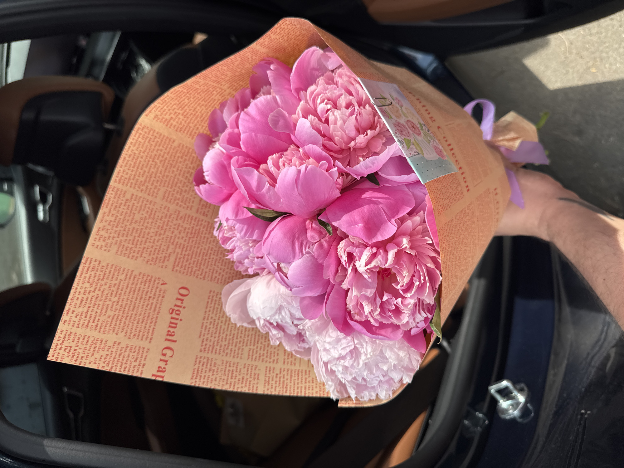 This media file features a close-up, overhead shot of a bouquet of vibrant pink peonies being held, likely within the interior of a vehicle. The peonies are the primary subject, showcasing their lush, layered petals in various shades of pink, some with lighter, almost white tips. They are wrapped in brown paper that has a printed text resembling old newspaper print, with the prominent phrase "Original Grap" visible in large red lettering. A small, decorative card is attached to the bouquet, along with a light purple ribbon tied around the stems, securing the wrapping.
The background suggests the setting is the dark interior of a car, with glimpses of leather seating and a black surface. Part of a person's arm and hand are visible on the right side, holding the bouquet, with some tattoos present. The lighting indicates it is daytime, likely sunny, casting shadows. The overall emotion conveyed is one of gifting and beauty, with the bright flowers standing out against the darker, enclosed space. The text on the wrapping paper is mostly illegible due to its fine print and the angle of the shot, but a few phrases and what appear to be names or headlines are partially visible, along with a title that appears to be "pning Collectio". The file may have been captured in Almaty, Kazakhstan, based on the provided location context, though no specific visual cues within the image confirm this.