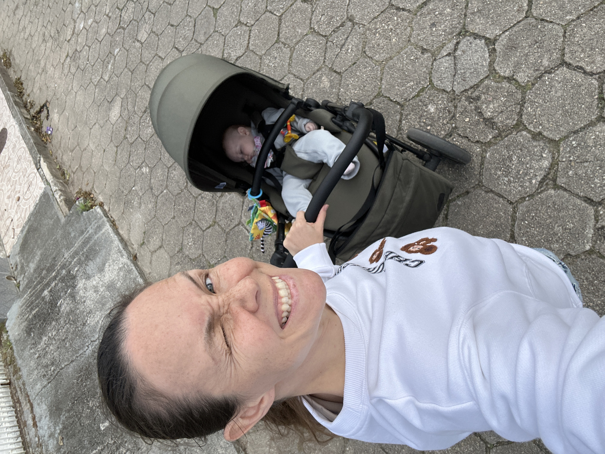 A woman with brown hair pulled back in a ponytail is taking a selfie outdoors, winking and smiling widely at the camera. She is wearing a white sweatshirt with embroidered designs. Next to her, a baby is sleeping in a khaki-colored stroller. The ground is paved with hexagonal concrete stones, and there is a concrete sidewalk with some grass and leaves in the background. The scene appears to be a residential street in Florianópolis, Brazil.