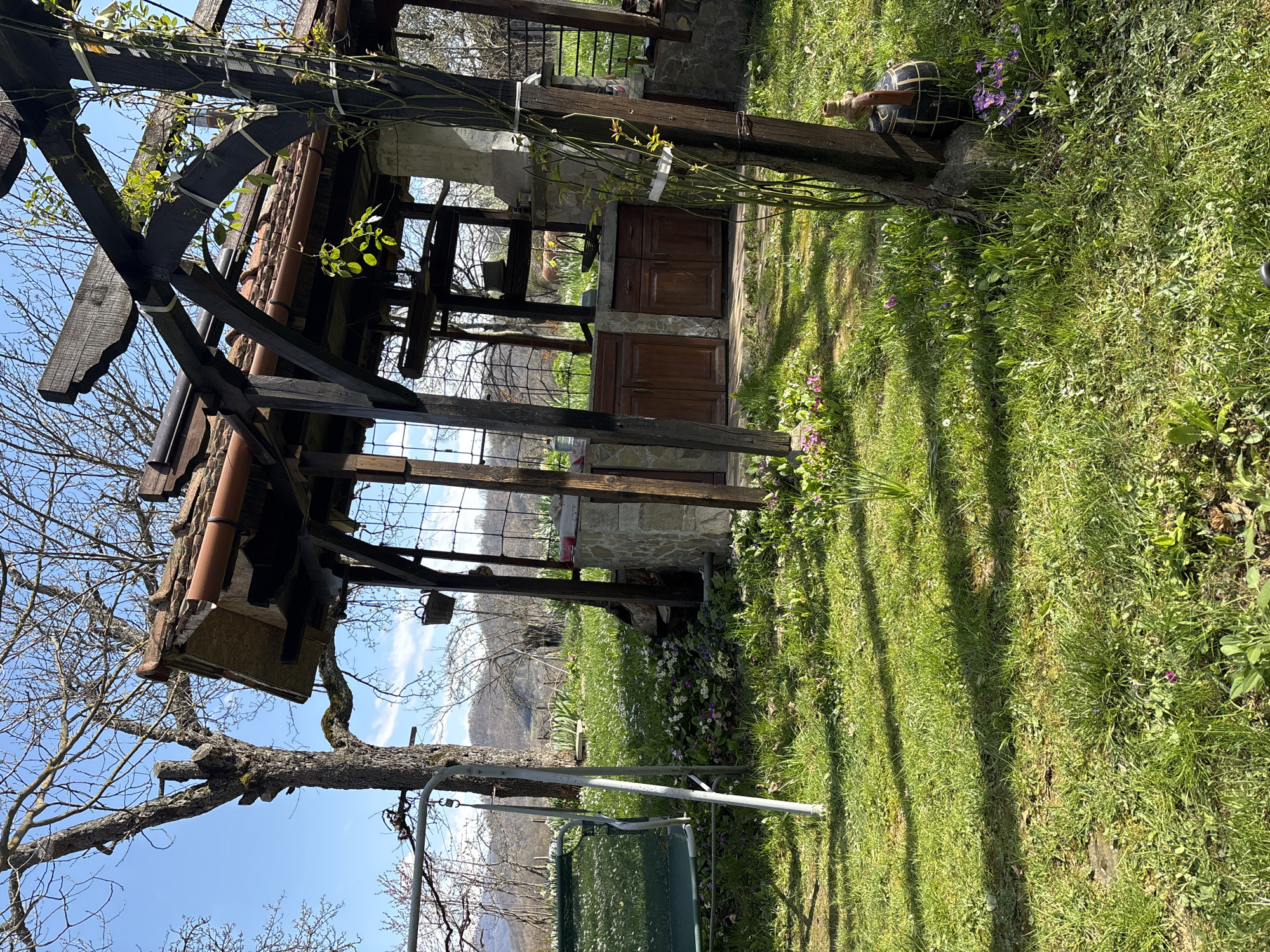 The image shows a partially constructed or rustic outdoor kitchen structure in a grassy yard. The structure features dark, heavy timber framing and a tiled roof. It has open sides with a window-like grid and a set of wooden cabinet doors set into a stone base. To the left, a bare tree branch extends into the frame, with a swing set hanging from it. In the background are distant hills or mountains under a blue sky. The yard is green with patches of flowers. A decorative wooden barrel sits in the grass next to the structure. The location is in Gorno Trape, Bulgaria.