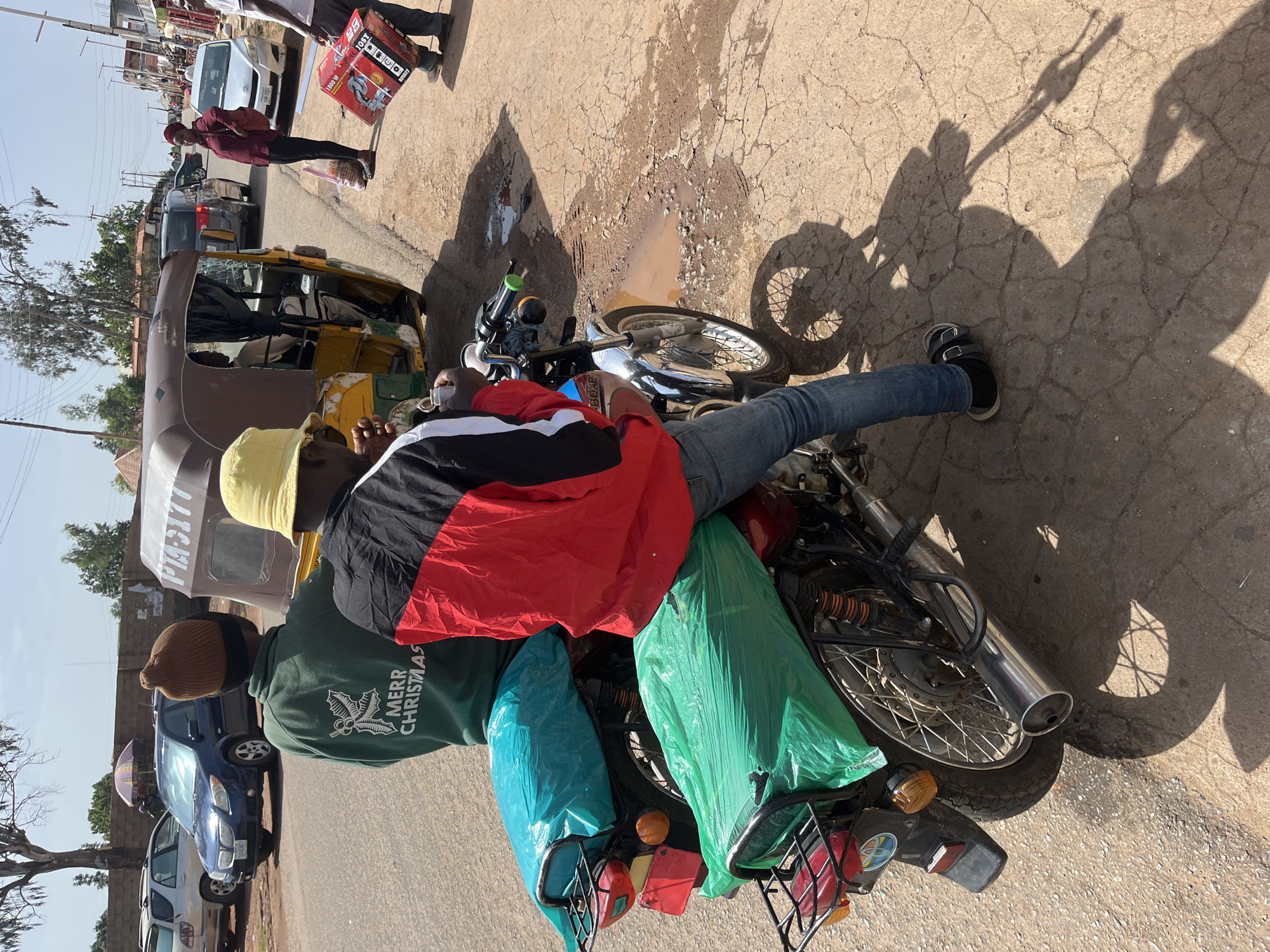The image depicts a street scene in Gura Top, Nigeria, with two individuals seated on a motorcycle. The motorcycle has two seats, both covered with green plastic sheeting. The person in the foreground is wearing a red, black, and white jacket and blue jeans. They are holding a small white object. The person behind them is wearing a green hooded sweatshirt with "MERR CHRISTMAS" printed on the back in white text. A yellow bucket hat is worn by the person in the foreground. In the background, a yellow and brown auto-rickshaw is visible, along with other vehicles and buildings lining the street. The ground is paved with cracked asphalt, and there are puddles of water in some areas. The sky is clear and the sun is shining, casting shadows on the ground. A person is seen walking in the background carrying a red box.