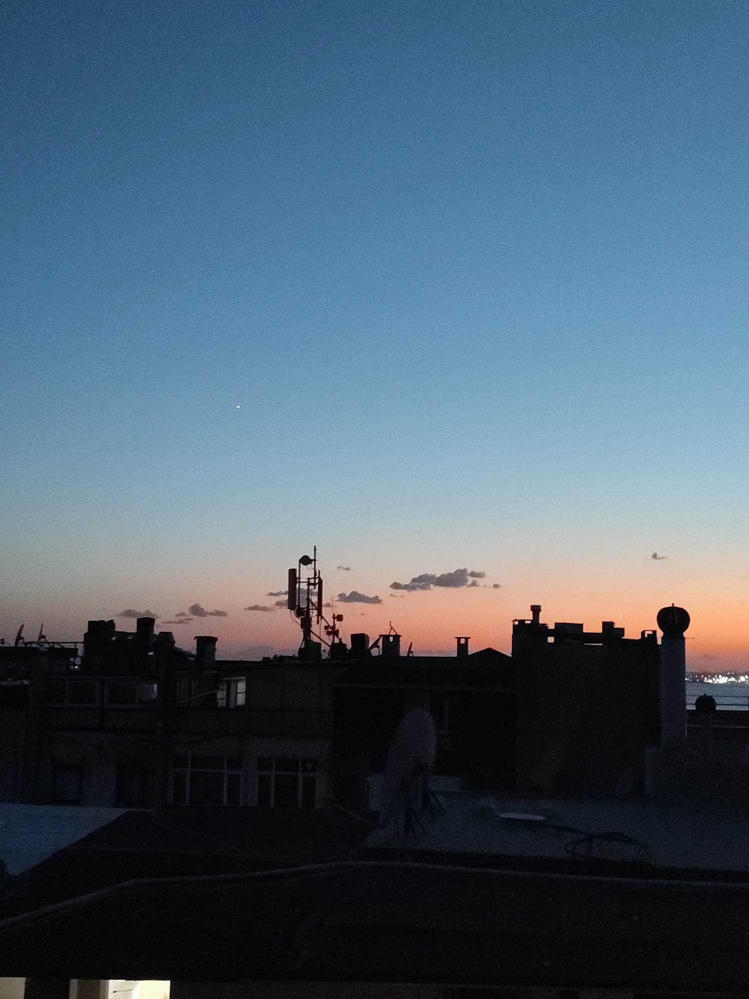 The image captures a twilight sky over the cityscape of Kadıköy, Turkey. The sky transitions from a deep blue at the top to a pale orange and pink hue near the horizon. Silhouetted against this sky are the rooftops of buildings, characterized by their dark, indistinct shapes. Prominent structures include communication towers with multiple antennas and parabolic dishes, indicative of urban infrastructure.
Several buildings are visible, some with visible windows and architectural details obscured by the dim light. A crescent moon is visible in the upper right portion of the sky. Small, scattered clouds drift across the horizon. The foreground consists of darker, lower rooftops, suggesting a layered urban environment. The overall impression is one of a quiet evening in a densely populated area.