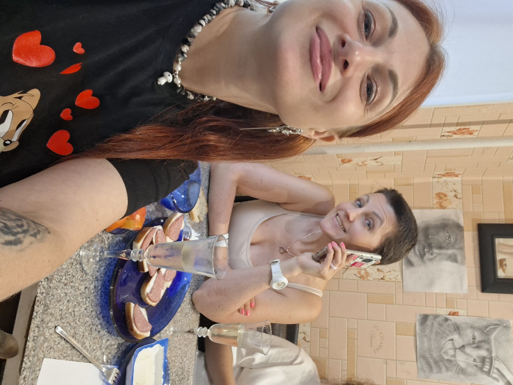 A selfie is taken by a woman with reddish-brown hair, wearing a black t-shirt adorned with red hearts and a mushroom pendant necklace. She is smiling and looking at the camera. In the background, another woman with short dark hair is holding a smartphone to her ear and smiling. She is wearing a light-colored tank top and a white wristwatch. On the table in front of them are several glasses of champagne, a plate of open-faced sandwiches with sliced meat, and a blue bowl. The setting appears to be indoors, possibly a kitchen or dining area, with tiled walls and framed drawings on the wall, one of which is a portrait of a cat and another of a crying child. There are no visible landmarks specific to Myadzyel, Belarus, in the image.