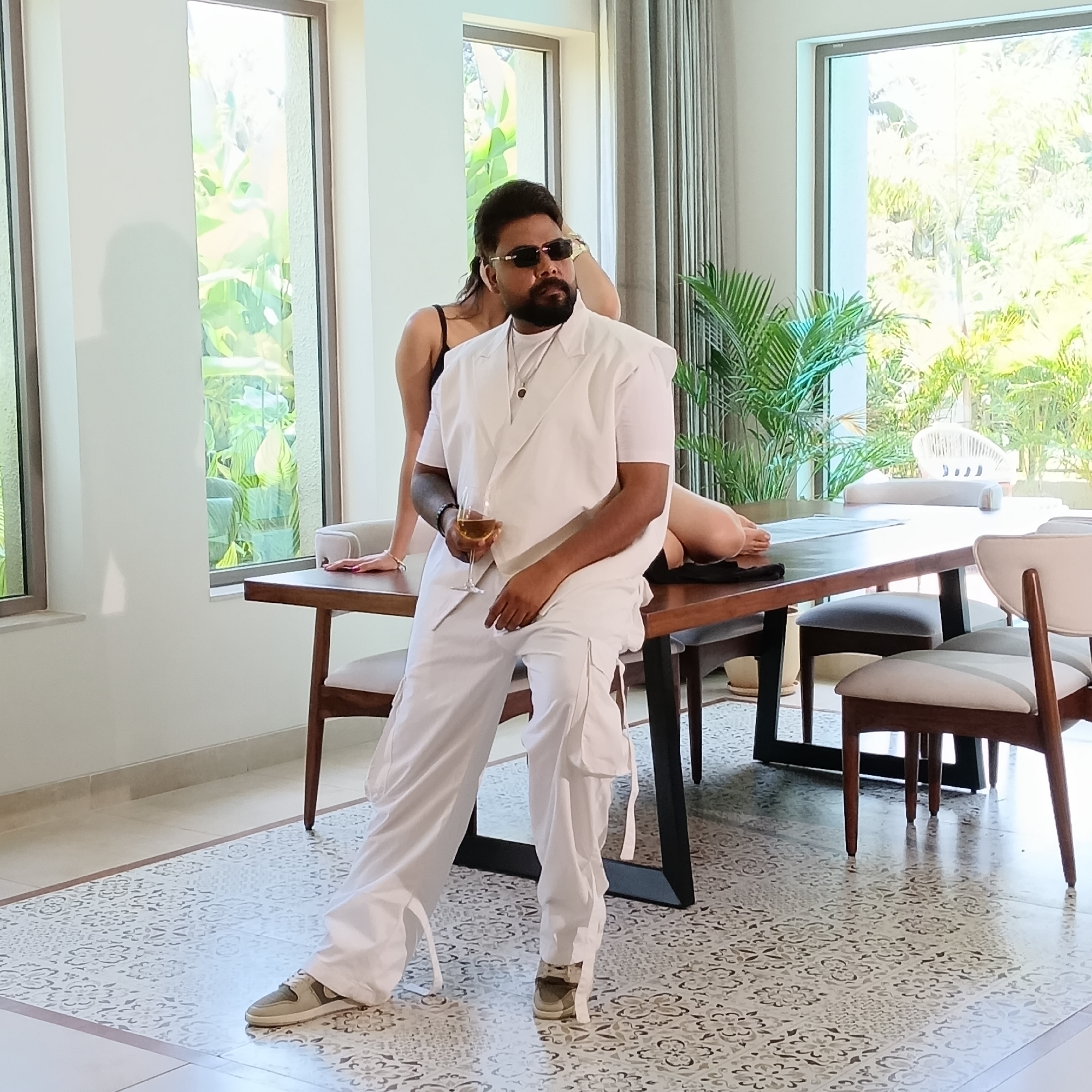 The image shows a man in a white outfit, sunglasses, and shoes, holding a wine glass while sitting on a dark wood table. A woman is partially visible behind him, lying on the table. They are in a room with large windows revealing outdoor greenery. The interior has a decorative tile floor, white walls, and chairs around the table. The location is likely Ashvem, India, based on visual cues such as architectural style and vegetation.