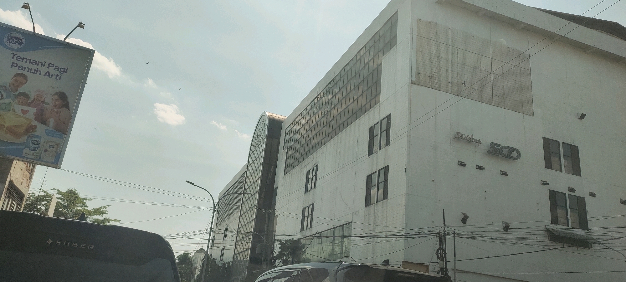 The image shows a white building with multiple floors, featuring a large arched glass section on one side and numerous rectangular windows. The building appears to be a commercial or office space, possibly a hotel or a department store, given the signage that includes "SCD" and what appears to be "BabyShop". The building is located in Semarang, Indonesia, as suggested by the presence of an advertisement for "Frisian Flag" milk, a popular brand in Indonesia. The advertisement features a family and the tagline "Temani Pagi Penuh Arti," which translates to "Accompany the Morning Full of Meaning." The environment is urban, with visible power lines crisscrossing the scene. In the foreground, the top of a dark-colored vehicle is visible, with the word "SABER" printed on it. The sky is mostly clear with a few scattered clouds, and the overall lighting suggests it is daytime.
