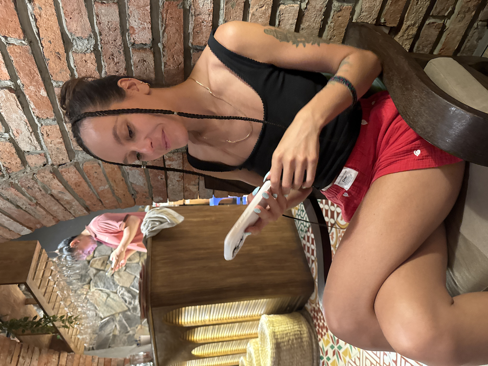 A woman with braided hair sits in a chair, looking at a smartphone in her hands. She wears a black tank top and red shorts. She is in a dimly lit indoor setting with exposed brick walls and patterned floor tiles. In the background, a man in a pink shirt is visible, also looking at a phone. A wooden bar counter with glasses hanging above it is also present. The overall ambiance suggests a casual establishment in Hà Nội, Vietnam.