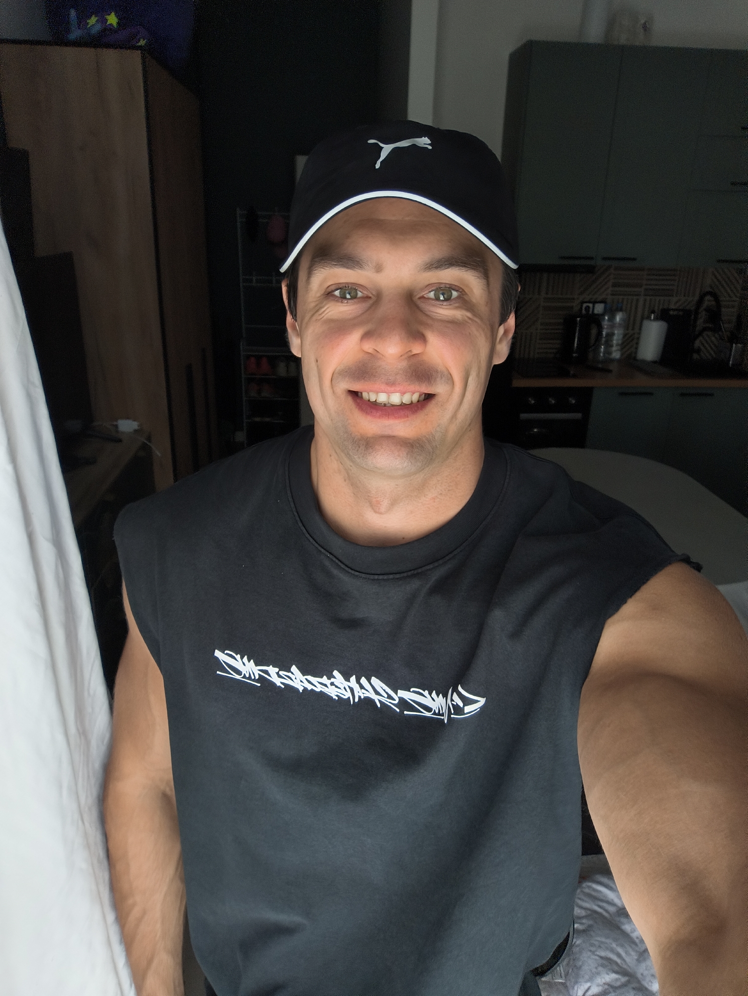 A man with light brown hair and green eyes is taking a selfie indoors. He is wearing a black baseball cap with a white Puma logo and a black sleeveless t-shirt with white graphic text. He is smiling at the camera, showing his teeth. The background is out of focus but appears to be a room with kitchen cabinets and a countertop visible. There is also a large piece of furniture, possibly a wardrobe or closet, to the left of the man. The overall lighting is dim, suggesting it might be evening or a room with limited natural light. The image does not contain any recognizable landmarks or elements that would place it specifically in Moscow, Russia.