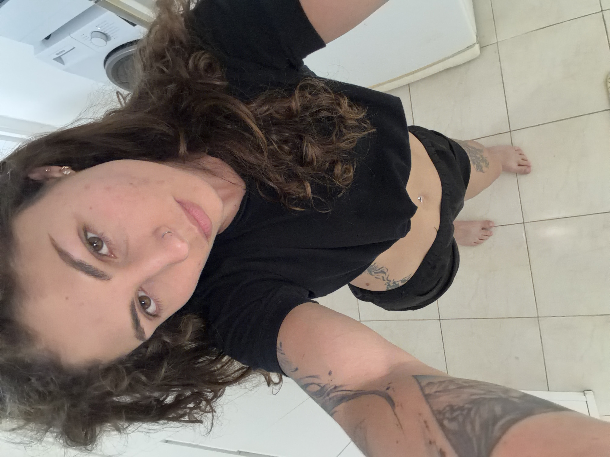 A woman with curly brown hair is taking a selfie in a room with light-colored tiled flooring. She is wearing a short black t-shirt and black shorts. Tattoos are visible on her arm, leg, and torso. A washing machine is partially visible in the upper left corner of the frame. The location is likely a private residence, but there is no additional information to confirm the location in the city of Kiryat Bialik, Israel.