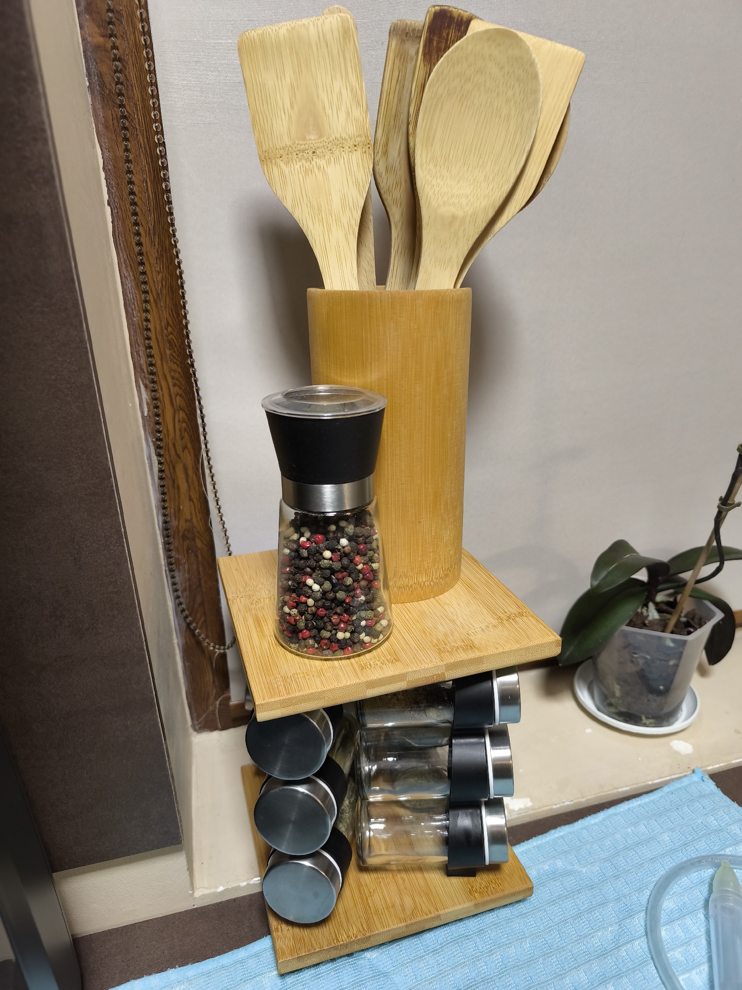 Here is a forensic description of the image:
**Scene:** Interior kitchen setting, location Unknown, Unknown.
**Objects:**
* A bamboo utensil holder filled with wooden spoons and spatulas.
* A pepper grinder filled with peppercorns placed on a bamboo shelf.
* A spice rack with several glass spice jars with metallic lids on a bamboo stand.
* A potted orchid plant with green leaves and exposed roots in a clear plastic pot, sitting on a white saucer.
* A light blue dish towel
* A clear plastic baby aspirator.
* Window with a dark brown frame and chain
**Environment:** Neutral colored wall. Close proximity to dark brown cabinetry or wall panel. Limited natural light.
**Details:**
* The spice rack appears to be multi-tiered and constructed of bamboo.
* The peppercorns in the grinder are a mix of red, black, white, and green.
* The wooden utensils in the holder appear to be new and unused.
* The orchid pot is clear, and the plant appears to be healthy.
* The scene is organized and tidy.
