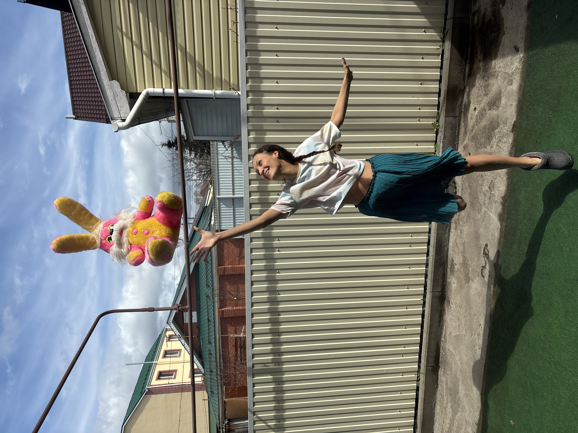 A woman is suspended horizontally in the air, smiling and reaching toward a large stuffed rabbit hanging from a metal rod. The woman is wearing a tie-dye shirt and a teal skirt, with grey footwear on her extended leg. The scene takes place outdoors against a backdrop of houses with various siding materials (yellow, brown, and green). A metal fence is partially visible behind the woman. The sky is blue with some clouds. The location is Ulyanovsk, Russia.