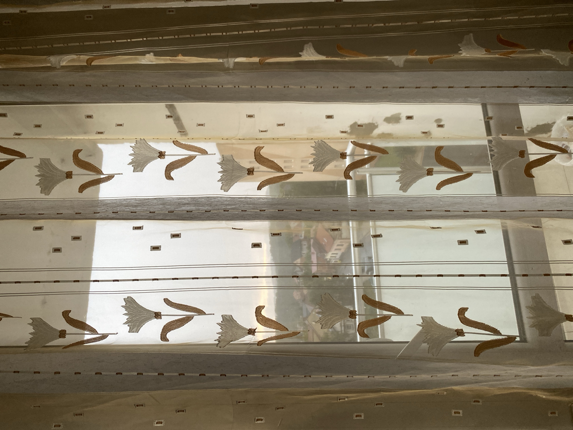 This is a photograph taken from inside an apartment in Bílina, Czechia, looking out through a sheer curtain. The curtain has a repeating pattern of white flowers with brown stems and leaves, and small brown squares. The window looks out onto a residential area with buildings, rooftops, trees, and a balcony. The sky appears overcast. There are no people visible in the image.
