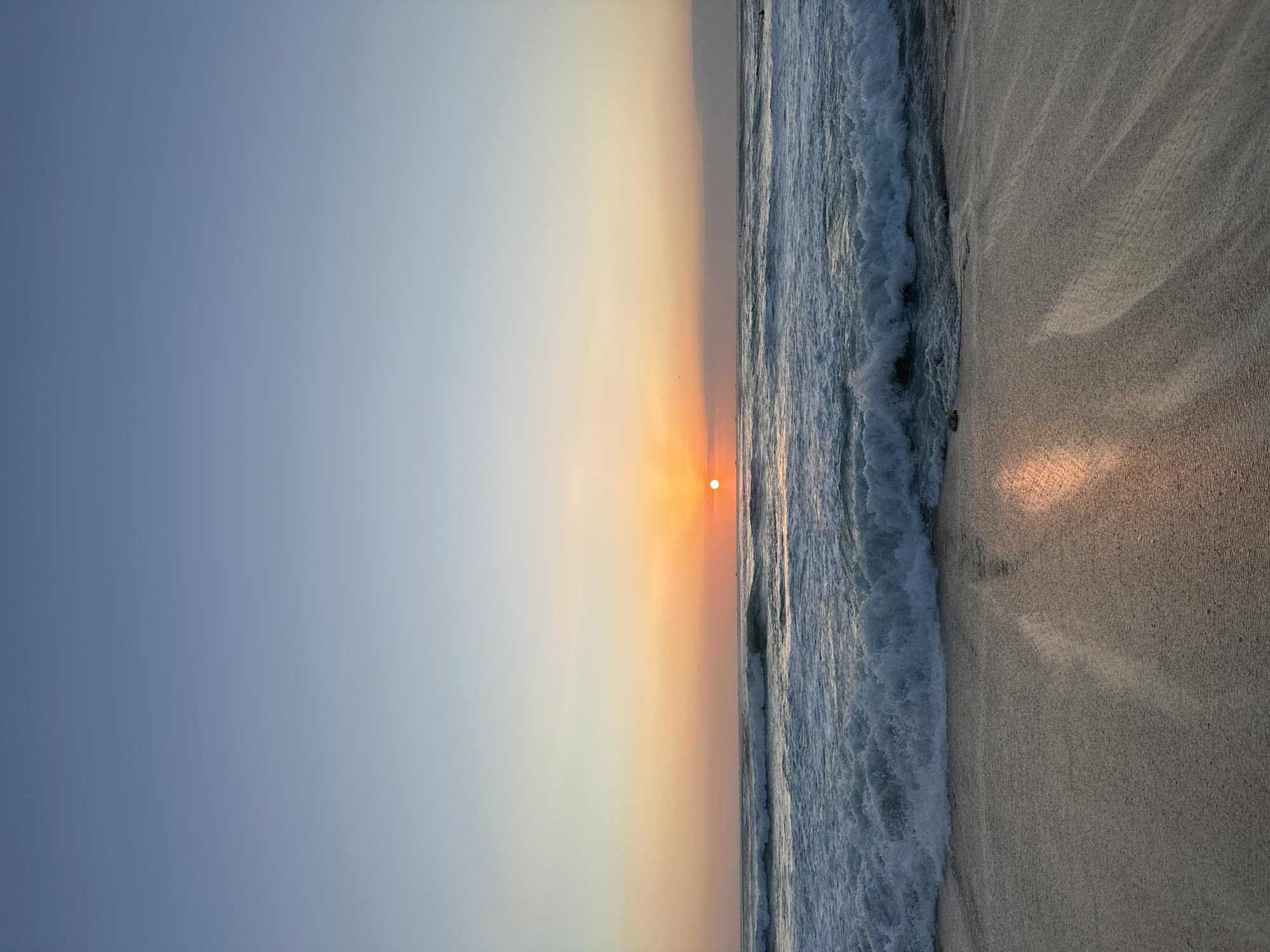 The image depicts a serene beach scene at sunset in Kuta Selatan, Indonesia. The sky transitions from a clear, pale blue at the top to a warm gradient of orange and yellow near the horizon, where the sun is setting. The sun itself appears as a small, bright orange circle, positioned slightly to the left of the frame's center.
The ocean is active, with numerous waves rolling towards the shore. The waves are white-capped and appear to be of moderate size, creating a textured pattern across the water. The water's surface reflects the colors of the sunset, particularly in the wet sand at the edge of the beach.
The foreground consists of damp sand, showing the imprints of receding waves and some scattered debris, possibly small stones or shells. The light from the setting sun casts a warm glow on the sand, highlighting its texture. In the distance, on the horizon, there are faint silhouettes of what appear to be small boats or surfers, adding a sense of scale and activity to the otherwise tranquil scene. The overall atmosphere is peaceful and tranquil, characteristic of a beach at dusk.