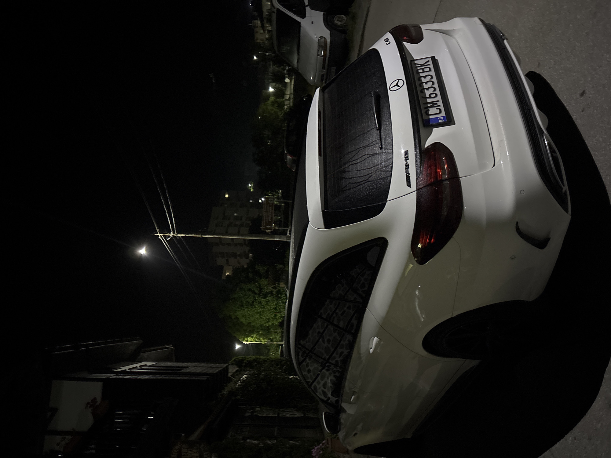 The image depicts a nighttime urban scene in Zlatograd, Bulgaria. The primary subject is a white Mercedes-Benz C43 AMG Estate, identified by its badging and wagon body style. The vehicle is parked facing away from the camera, slightly angled to the left, and bears a Bulgarian license plate "CM 6333 BK". Visible water streaks are present on its rear window, suggesting recent rain or heavy condensation.
To the immediate left of the Mercedes is a multi-story building with a dark facade, partially covered by green climbing foliage. A streetlight illuminates this area. In the background, multiple buildings are visible, including a taller structure on the left. A prominent sign in the mid-background reads "ДОБРЕ ДОШЛИ В ЗЛАТОГРАД ОБЩИНА ЗЛАТОГРАД" (Welcome to Zlatograd Municipality Zlatograd), confirming the specific location.
Further in the background, to the right, a white panel van (resembling a Renault Master) is parked, facing the camera. A dark-colored car is partially visible between the Mercedes and the white van. Overhead utility lines and a utility pole are visible on the right side of the frame. The scene is illuminated by multiple artificial light sources, casting shadows and highlighting various elements. No individuals are present in the image.