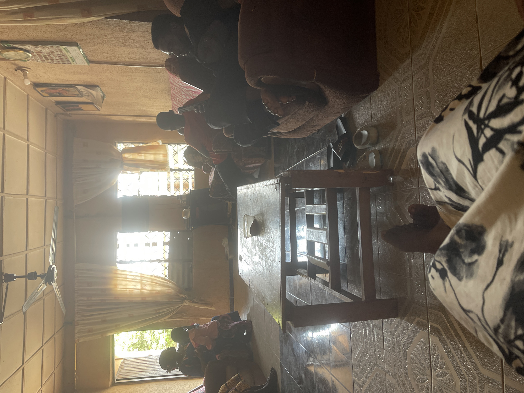 The image shows an interior scene in Sabon Tasha, Nigeria. A group of people are seated on what appear to be couches or chairs, mostly facing away from the camera. A wooden table is prominently positioned in the foreground. The room is brightly lit by natural light streaming through windows partially covered with curtains. A ceiling fan is visible. The floor is tiled with a patterned design. Several pairs of shoes sit on the floor near the table. A calendar and other small objects are affixed to the wall. A person's legs, adorned in black and white patterned clothing, are partially visible in the foreground.
