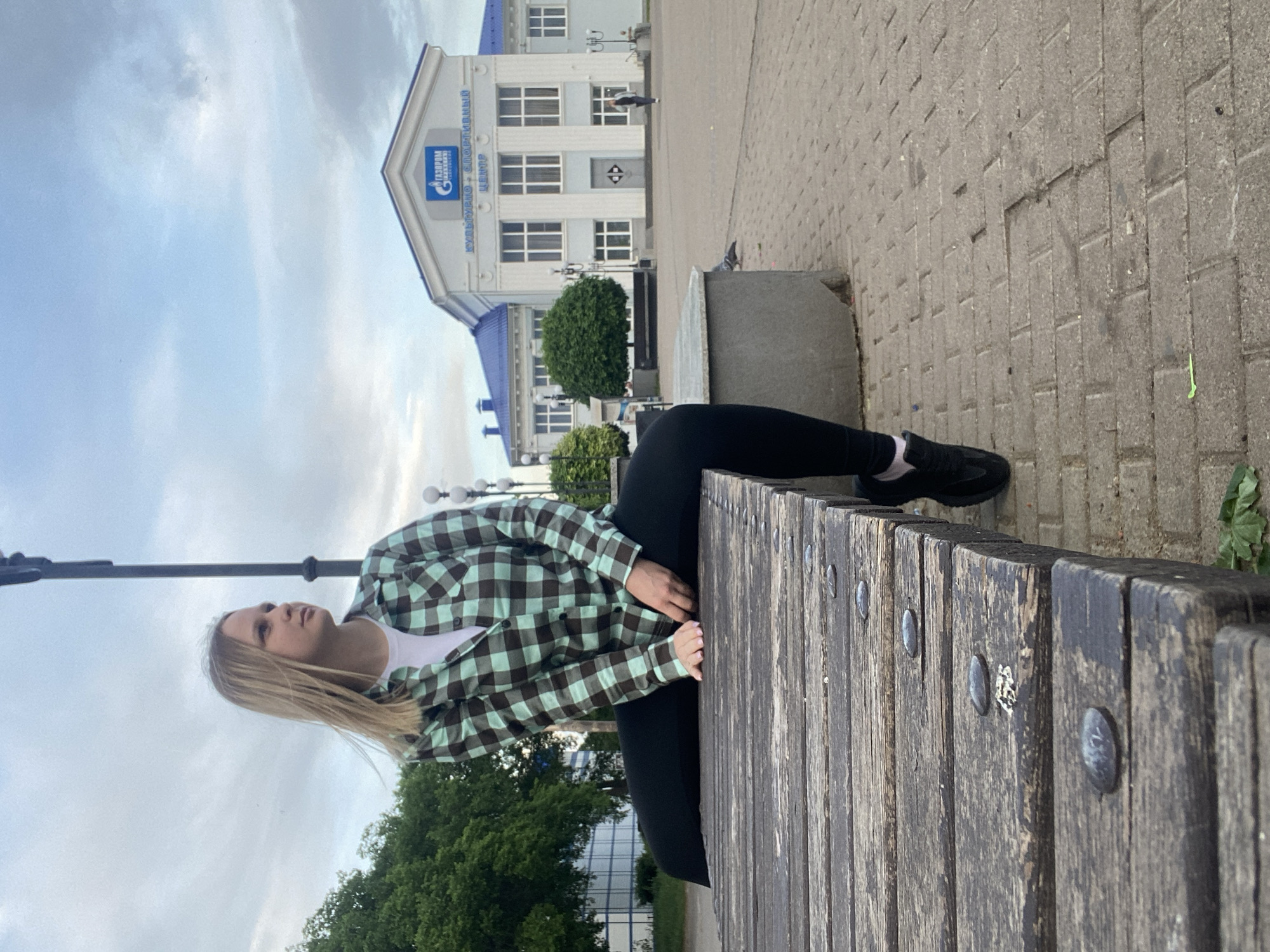 A young woman sits on a wooden bench in Chaikovsky, Russia. She wears black leggings, a white t-shirt, a plaid green and brown flannel, and black sneakers with white socks. She rests her hands on the bench. Behind her is a "Gazprom" cultural and sports center building. The sky is overcast. The bench is made of weathered planks with metal fasteners. A section of paved sidewalk is also visible.