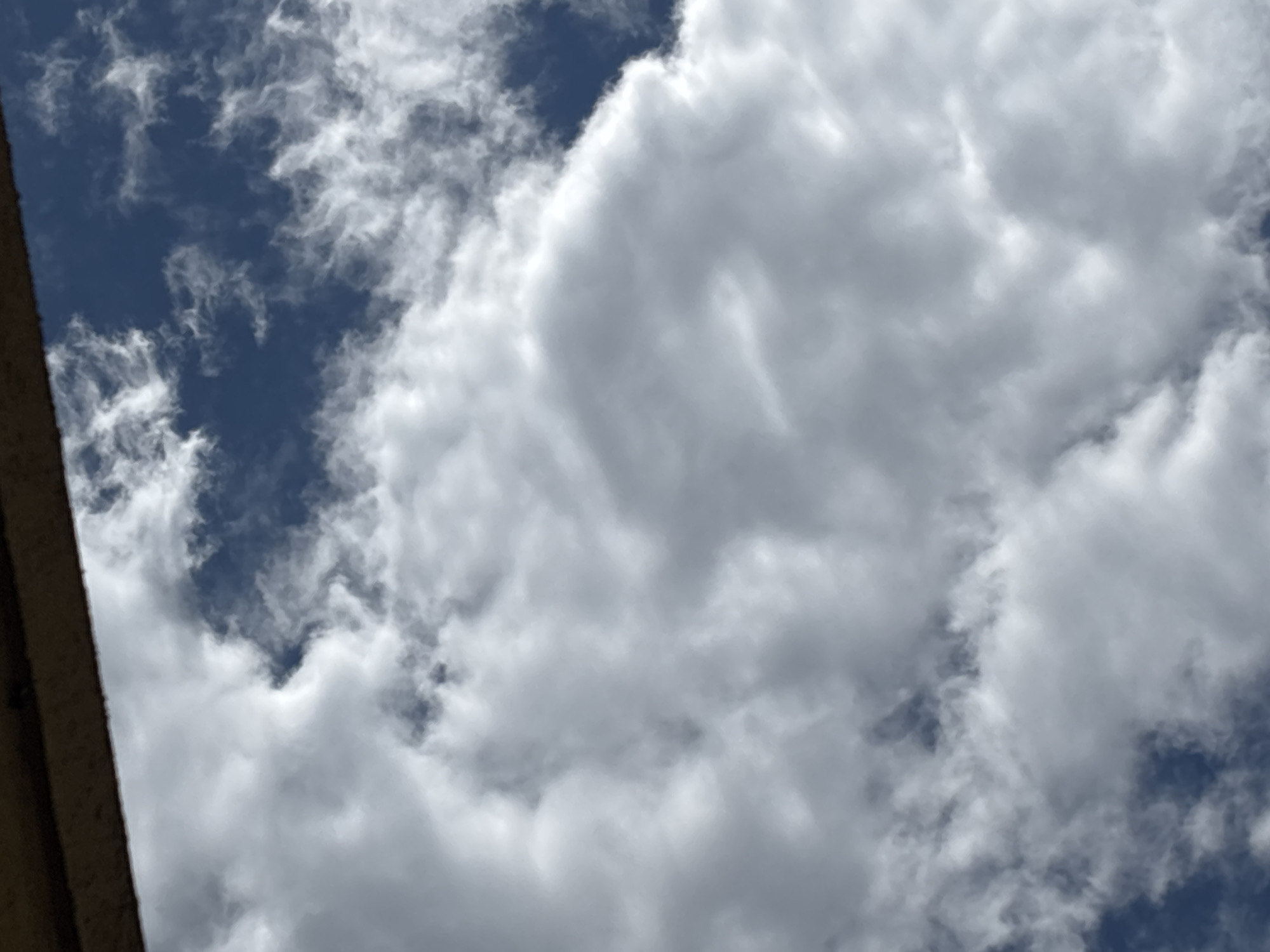 The image shows a view looking upwards into a sky filled with white and gray clouds, with patches of blue sky visible between the clouds. A portion of a building or structure is visible in the lower left corner of the image. The specific location in the city of Sofia, Bulgaria cannot be determined from this image alone.