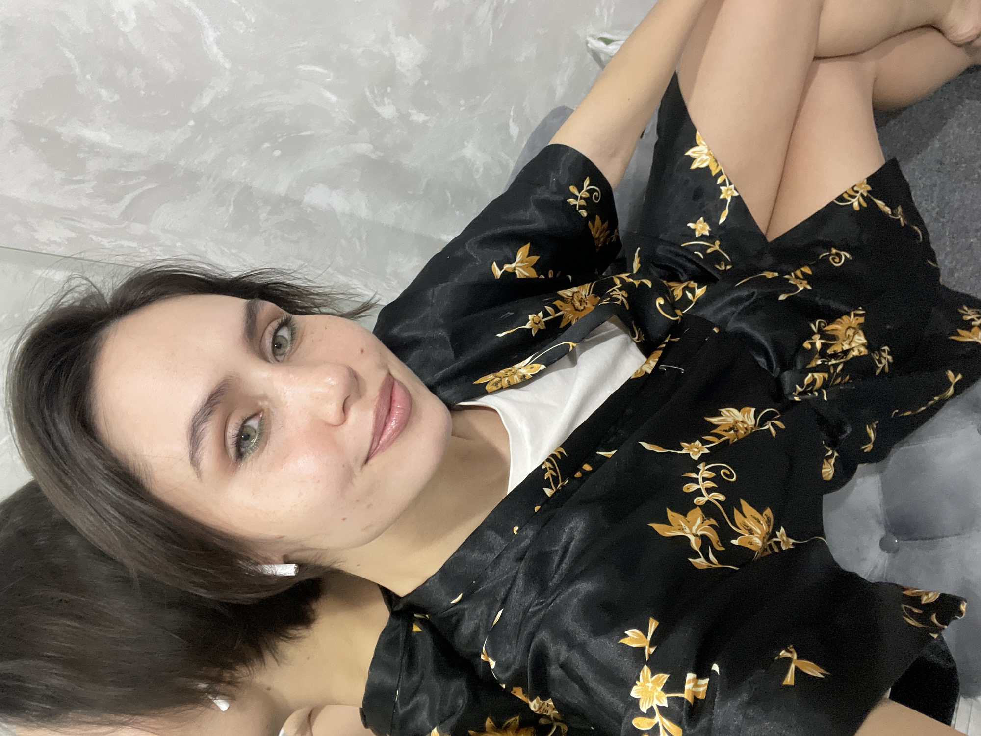A female individual is depicted in a self-portrait taken indoors in Izhevsk, Russia. She has short, dark hair, green eyes, a light complexion with visible freckles, and is wearing light makeup including green eyeshadow and pink lip gloss. She wears white, rectangular stud earrings. Her attire consists of a black, short-sleeved robe with golden floral patterns, worn open over a white t-shirt. She is positioned in a seated or reclining posture, with her legs crossed and partially visible. She is seated on a grey, upholstered surface. A reflective surface, likely a mirror, behind her shows a blurred reflection of her back and the side of her head. The wall visible beyond the reflective surface is grey with a discernible textured finish. No direct interactions with other individuals or subjects are visible.