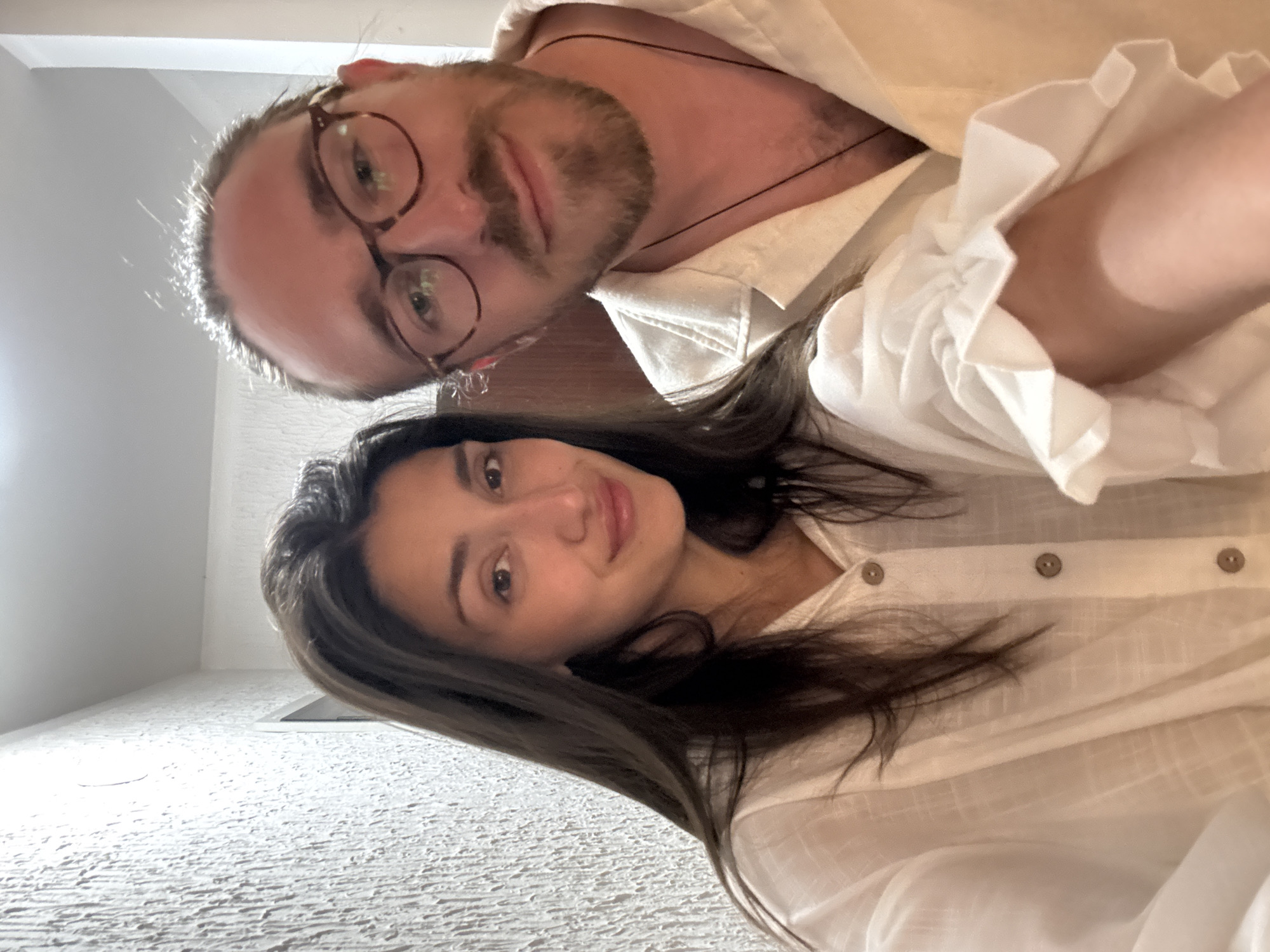 This is a close-up selfie-style portrait of two individuals, a man and a woman, positioned at a low angle looking upwards towards the camera. The image itself is rotated 90 degrees clockwise from its natural upright orientation.
The man, in the foreground on the left, has light brown hair, a beard, and a mustache. He wears distinctive round, brown-rimmed eyeglasses that reflect light. A thin, dark cord necklace is visible against his bare skin beneath his light-colored, collared shirt. The shirt, likely white or cream, features a subtle ruffled cuff detail on his sleeve.
Slightly behind him and to the right is the woman, who has long, dark brown hair that cascades over her shoulder. She is also wearing a light-colored, button-down shirt, with three brown buttons visible along the placket. Both subjects are looking directly at the camera with neutral to slightly smiling expressions, conveying a relaxed and pleasant mood.
The background consists of a textured, light-colored surface, possibly a wall or ceiling, indicating an indoor setting. The lighting is bright, with some backlighting apparent around the man's head. There is no visible text in the image. The scene suggests a casual moment captured between the two individuals. This media file is contextualized as being from Zankovići, Montenegro.