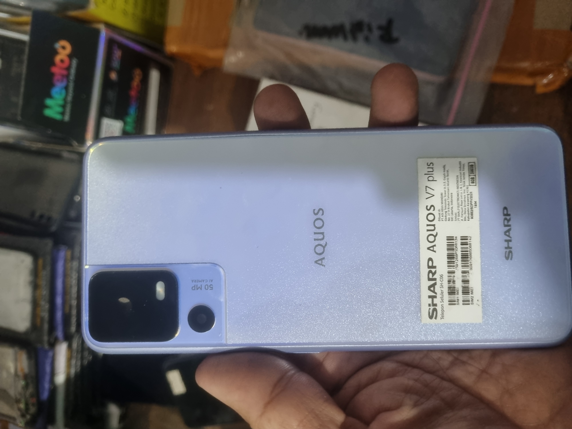 A light purple SHARP AQUOS V7 plus smartphone is held in a person's left hand, with the back of the phone facing the camera. The phone has a camera bump with two lenses, one large and one small, and text indicating "50 MP AI CAMERA". Below the camera, the brand name "AQUOS" is visible. A white sticker is present on the lower half of the phone, displaying "SHARP AQUOS V7 plus" and technical specifications including "SIM 1 IMEI" and "SIM 2 IMEI" with associated numbers. The text on the sticker also indicates that the product is manufactured in Indonesia by PT ADI REKA MANDIRI and PT SHARP ELECTRONICS INDONESIA, with addresses in Jakarta and Karawang respectively. The phone is positioned in front of a cluttered background of electronic components and packaging materials, suggesting a retail or repair environment. The lighting is artificial and appears to be from overhead sources. The image does not contain any information that would allow for determining its location in Probolinggo, Indonesia.