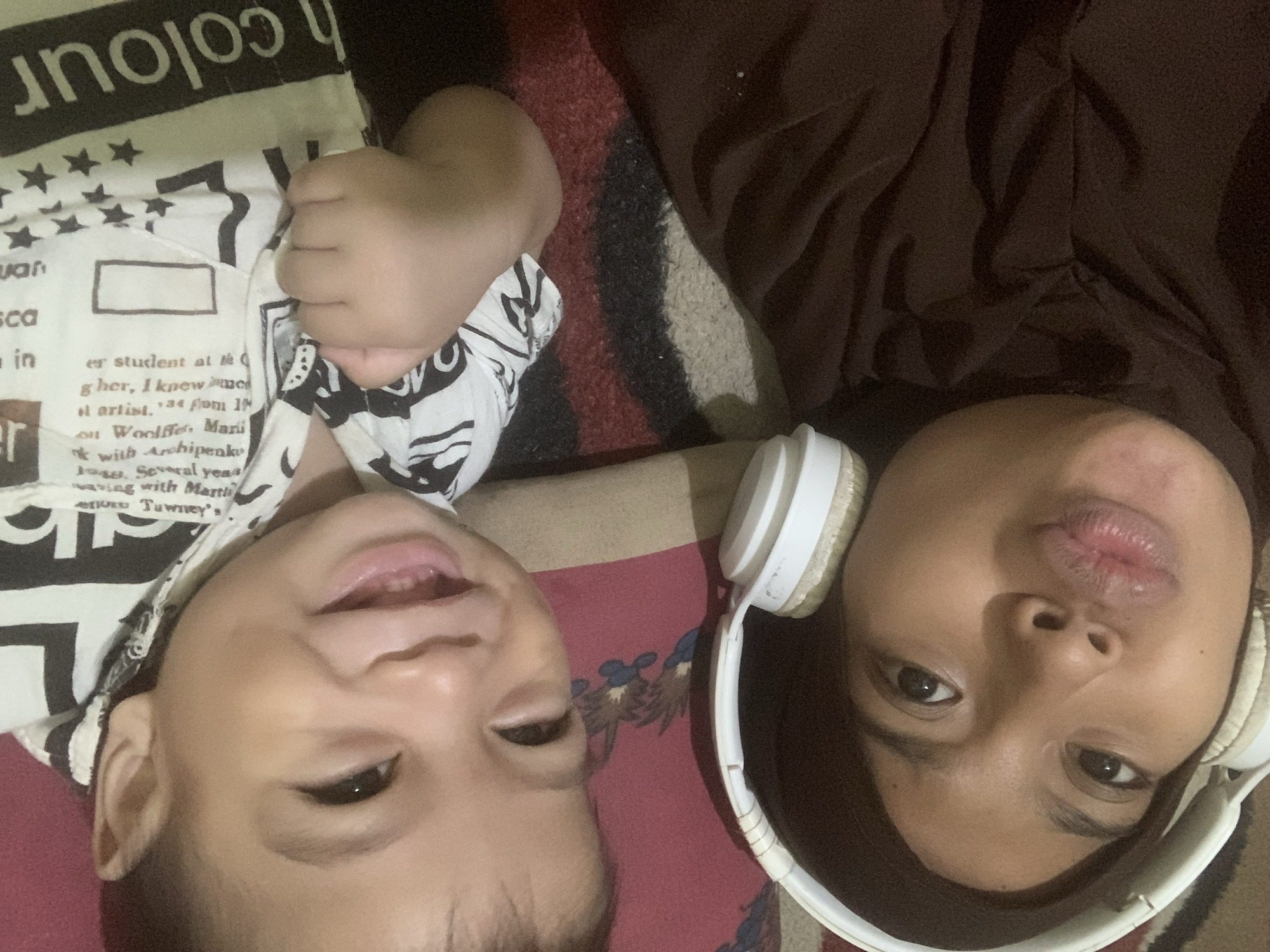 This media file is a close-up, overhead shot of two people lying down, their faces angled upwards. The person on the left appears to be a young child, with their mouth open and eyes looking upwards. They are wearing a white garment with black text and star patterns. The person on the right is wearing white headphones and has their eyes looking upwards, with their lips pursed. They are wearing a dark brown garment. The background appears to be a patterned rug. The lighting suggests it is indoors, possibly during the day or evening. Visible text on the child's garment includes partial words like "colour", "sca", "in", "er student at te", "gher, I knew me", "ol artist. 134 fom 10", "on Woolffer Mari", "with Archipenku", "1048. Several yeas", "wing with Martii", and "ende Tawney's". The overall impression is a casual, intimate moment captured between two individuals.