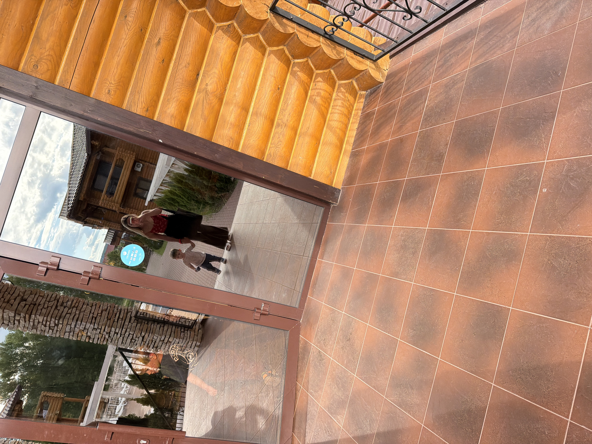 The image shows a woman holding a young child's hand, reflected in a large glass door. The woman is wearing a red top and dark pants, and the child is in a striped shirt and dark pants. They are standing on a tiled floor, with a wooden building visible in the reflection. To the right of the door, a large wooden structure with a balcony is visible. In the foreground, the tiled floor is a reddish-brown color. The overall scene appears to be an outdoor entrance to a building, possibly a resort or hotel, in Красный Ключ, Russia, given the context. The lighting suggests it is daytime, with sunlight casting shadows.