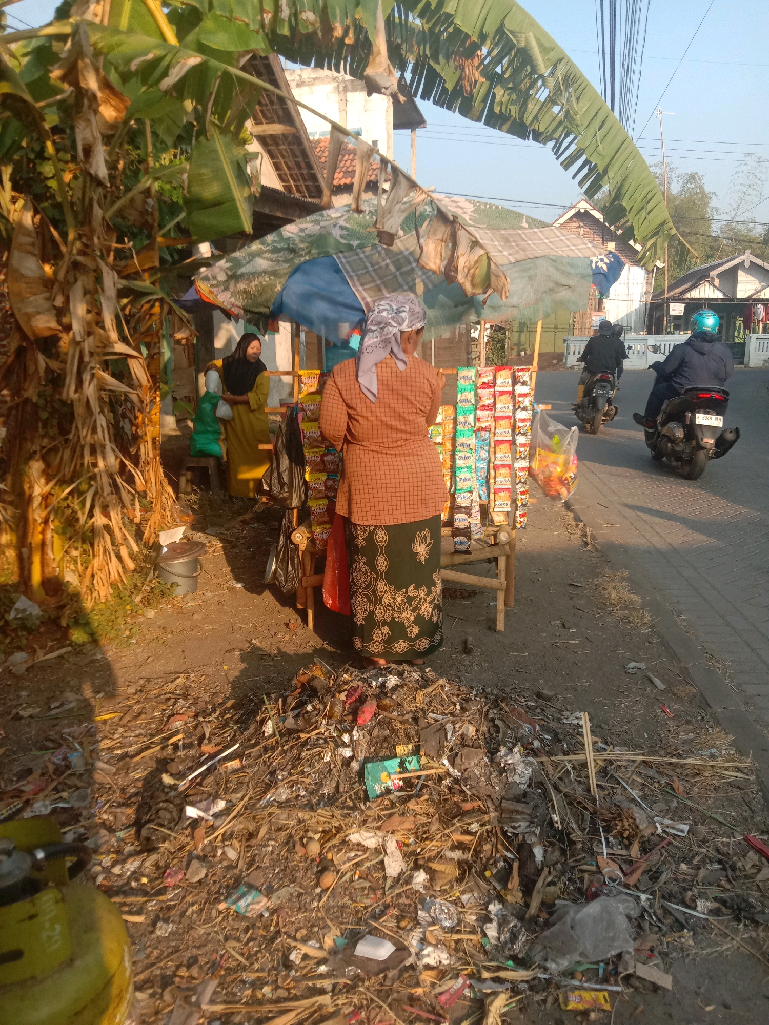 In Selotambak, Indonesia, a street vendor operates a small stall laden with snacks. The vendor, a woman in a patterned orange top and a green and black patterned skirt, stands behind the stall. To her left, another woman in a yellow dress and black hijab holds a green plastic bag. In the foreground, a large pile of diverse debris, including plastic wrappers, wood, and organic matter, is visible. Beyond the stall, two men on motorcycles pass by on the paved road. The scene is set against a backdrop of banana trees and residential buildings under a clear sky.