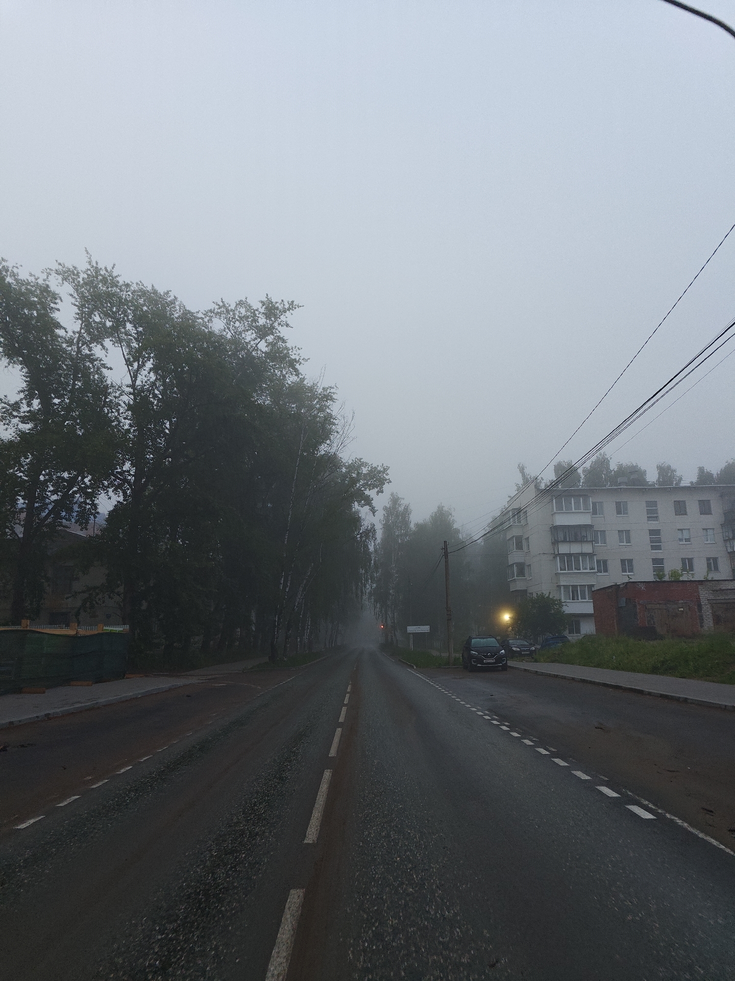 This image captures a street scene in Chaikovsky, Russia, heavily obscured by dense fog. A wet asphalt road, marked with white dashed lane lines, recedes into the misty distance, with visibility significantly limited.
On the left side of the road, a thick stand of tall deciduous trees, some appearing to be birches, forms a dark, green wall that gradually fades into the fog. A green construction fence is barely visible on the far left.
The right side of the street features a light-colored, multi-story apartment building with several balconies, its architectural details softened by the mist. Below it, a single glowing orange street light attempts to pierce the fog. A black Renault car is parked on the side of the road, partially visible, with its license plate `P354XX 159 RUS` identifying its origin from Perm Krai, Russia. Another indistinct vehicle is parked behind it. Further right, a low red brick building is situated amidst overgrown grass. Power lines stretch across the upper right section of the sky, disappearing into the pervasive haze.
No people are visible. The heavy fog and the illuminated street light suggest either early morning or late evening. The scene conveys a quiet, somewhat mysterious, and tranquil atmosphere due to the weather conditions and lack of activity.