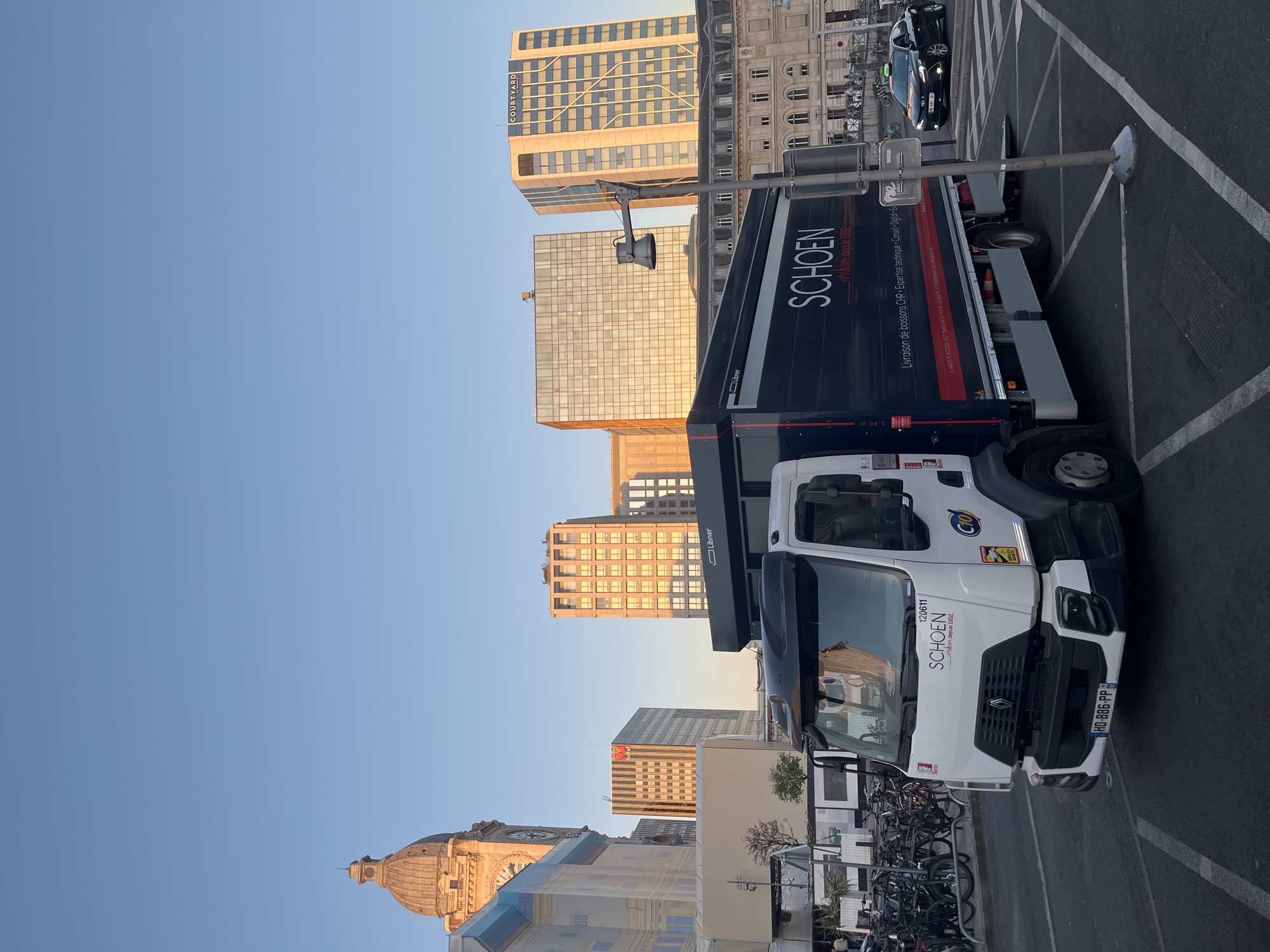 In Paris, France, at street level, a white truck with a dark cargo box is parked. The truck has the "SCHOEN" logo on its side, along with text indicating "Livraison de boissons CHR. Expertise technique. Conseil Digital. Event". The truck's license plate reads "HO-886-PP". To the right of the truck, a black sedan, possibly a taxi, is visible. In the background, several modern buildings rise, including one with a "COURTYARD" sign. A clock tower with a dome is also prominent. The sky is clear and blue. The scene appears to be a commercial or business district, possibly during the daytime with the golden hour sunlight illuminating some of the buildings.