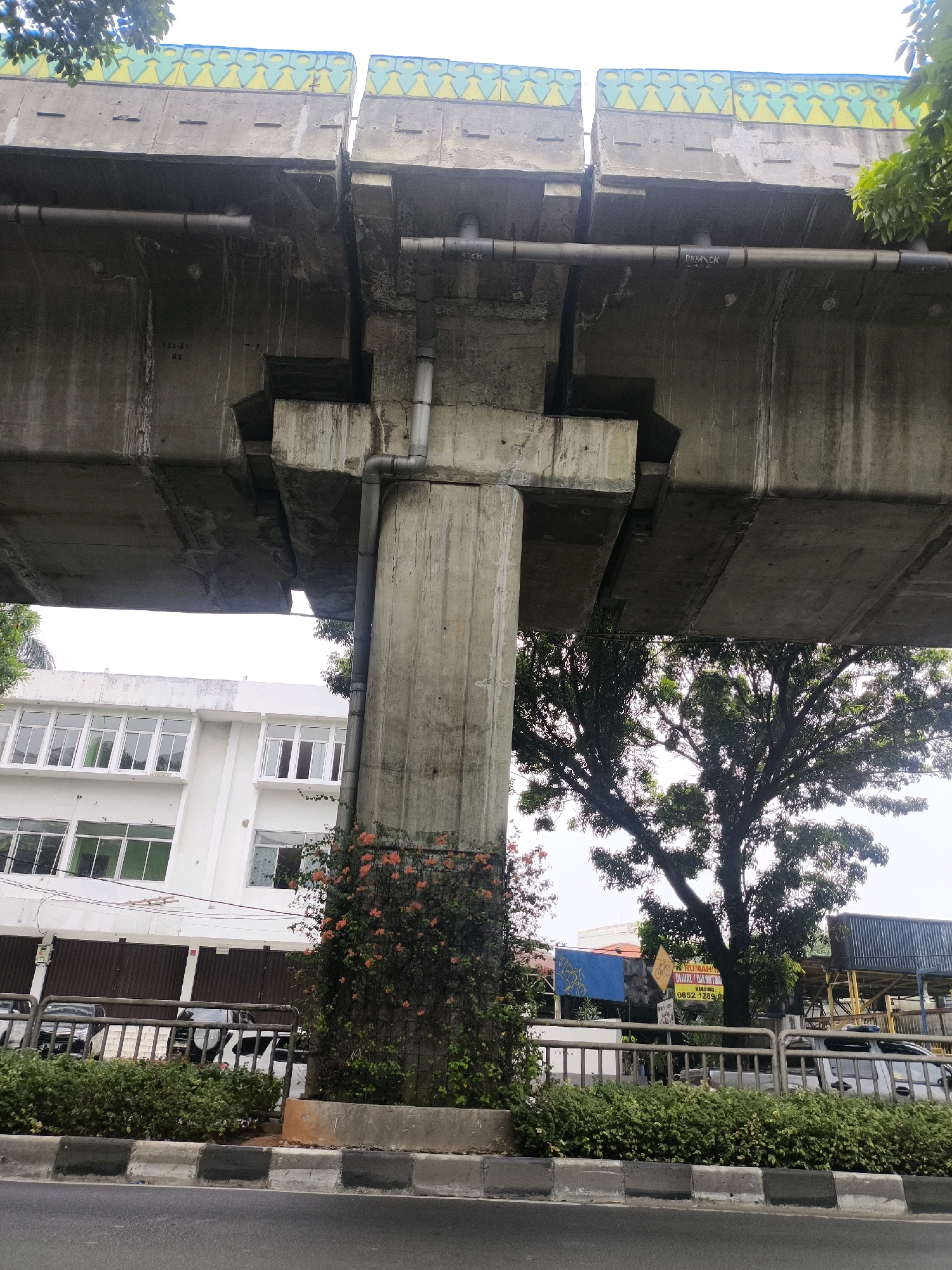 The image shows an elevated concrete structure, likely a bridge or overpass, with visible pipes running underneath. The concrete supports are massive, with one prominent pillar covered in orange flowering vines. To the left of the pillar is a modern white building with numerous windows, and to the right, a large tree with dense foliage. A metal fence and hedges separate the street from the area beneath the overpass. The street itself is paved, with a concrete curb and sidewalk. The sky is overcast. This scene is located in South Jakarta, Indonesia, likely on or near Jalan Gatot Subroto, a major thoroughfare known for its elevated infrastructure.