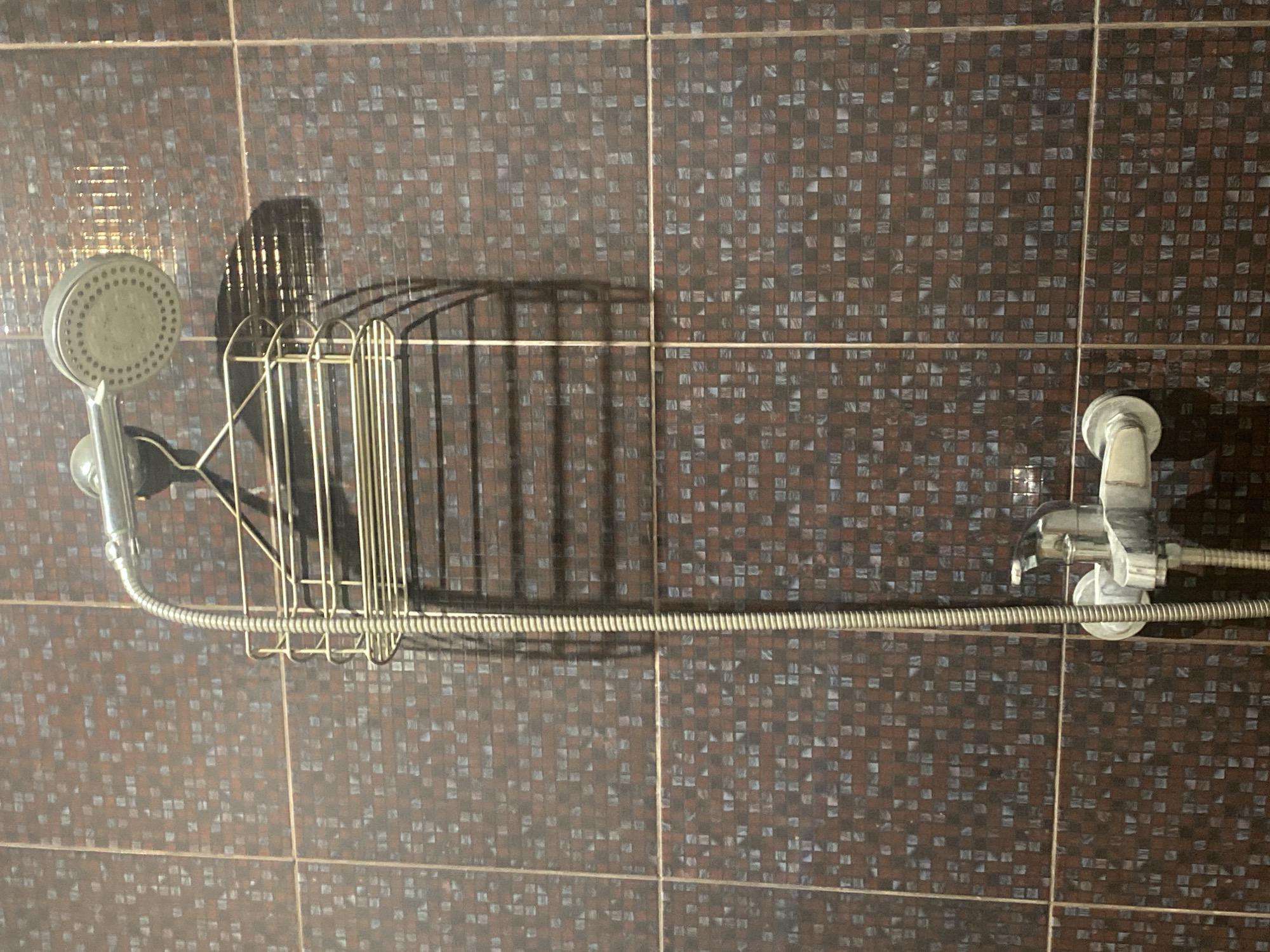 A close-up, waist-up shot shows a shower head, a metal shower caddy, and a faucet mounted on a tiled wall. The wall is covered in small, rectangular tiles with a dark brown and grey mosaic pattern. The shower head, made of polished metal, is positioned at the top of the frame, connected to a flexible hose. Below it, a gold-colored wire shower caddy hangs from the shower head. At the bottom of the frame, a metal faucet with white handles is visible. The scene appears to be in a bathroom, possibly in Chișinău, Moldova, given the context provided.