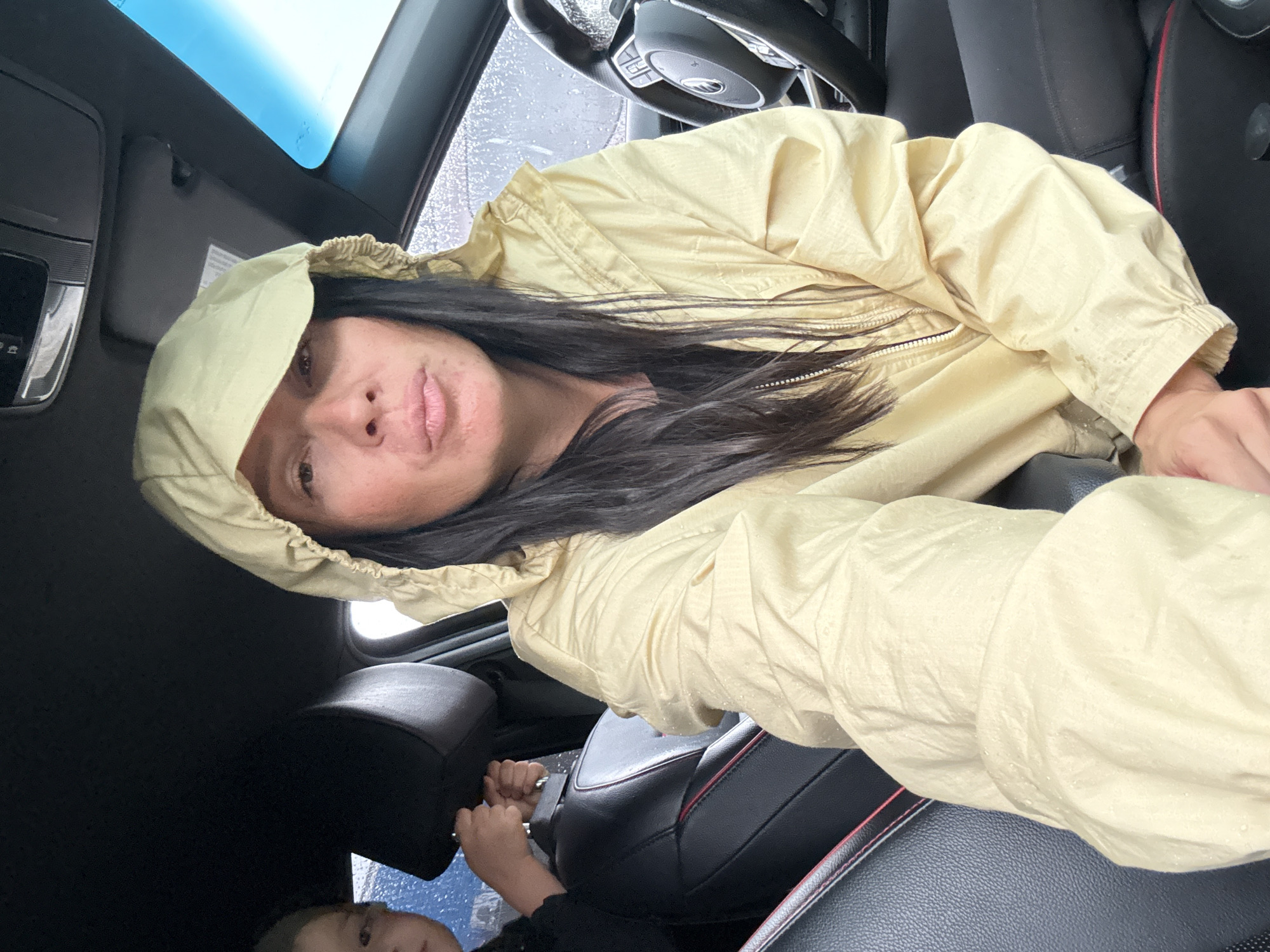 A selfie taken from the passenger seat of a vehicle shows a woman wearing a light yellow hooded jacket. The jacket appears to be water-repellent, with visible water droplets on the fabric. Her hair is dark and extends down past her shoulders. She is looking directly at the camera with her lips pursed. A young child is partially visible in the background to the left, seated in the back of the vehicle. The interior of the car is visible, including a black leather seat with red trim and a steering wheel. The sky outside the car windows appears overcast and rainy. The scene is likely taking place in the city of Chaikovsky, Russia, given the user's prompt.