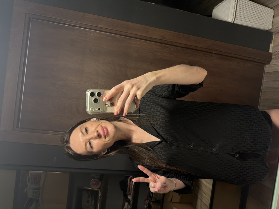 A woman poses in a mirror selfie, holding up a phone with a clear case. She wears a dark, patterned button-up shirt and gives a peace sign. She has long brown hair, fair skin, and is wearing an earpiece. The reflection shows a wardrobe with shelves containing various items. Behind her is a dark wooden door and a wicker basket on top of the wardrobe. The floor appears to be made of dark wood.