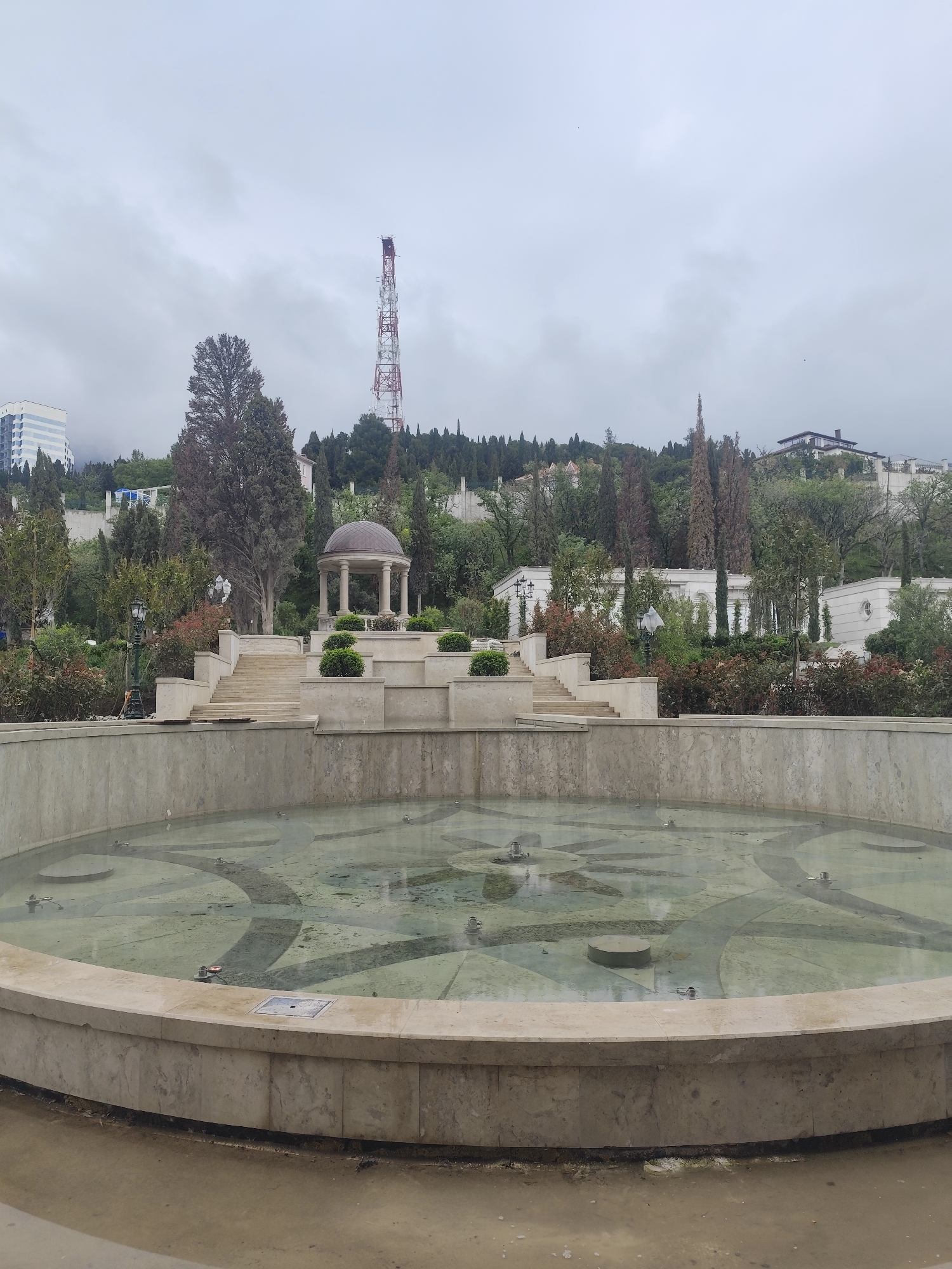 The photograph shows a concrete fountain in a park setting. The fountain is circular and contains a small amount of water with a visible design on the bottom. Behind the fountain is a tiered structure with stairs leading to a gazebo-like structure at the top. The surrounding landscape includes trees, shrubs, and various buildings on a hillside. A tall telecommunications tower is visible on the horizon. The sky is overcast. The location is in the city of Unknown, Unknown.