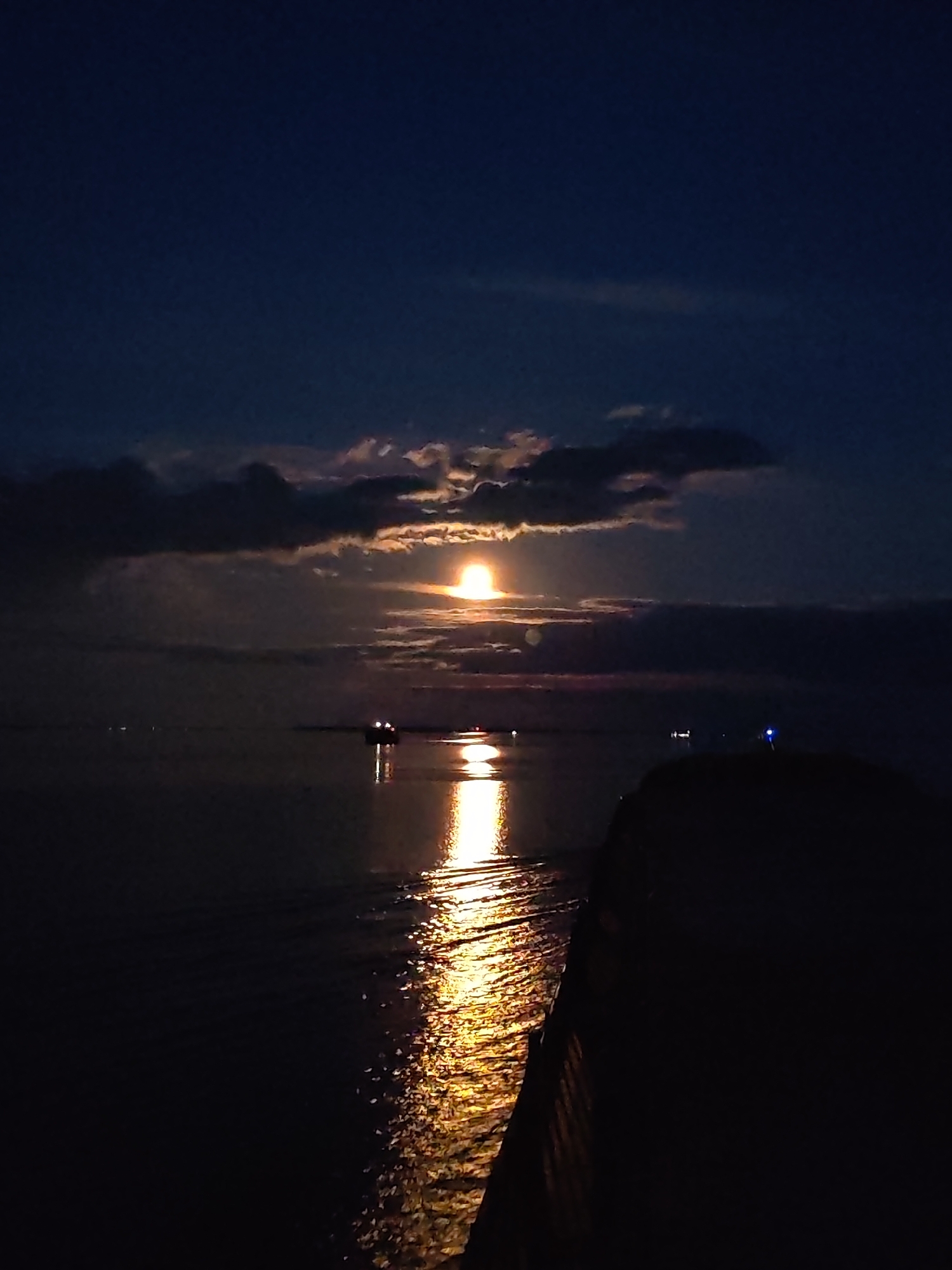 Captured in Bol'shie Mkhi, Russia, the scene presents a nocturnal view dominated by a body of water reflecting a bright, possibly full, moon. The moon is partially obscured by clouds, creating a diffused glow. A ship with visible lights is positioned on the water, its illumination mirrored in the water's surface. A dark landmass or structure is silhouetted in the foreground, obscuring the lower right portion of the frame. The sky is a gradient of dark blues, suggesting a late evening or early morning hour.