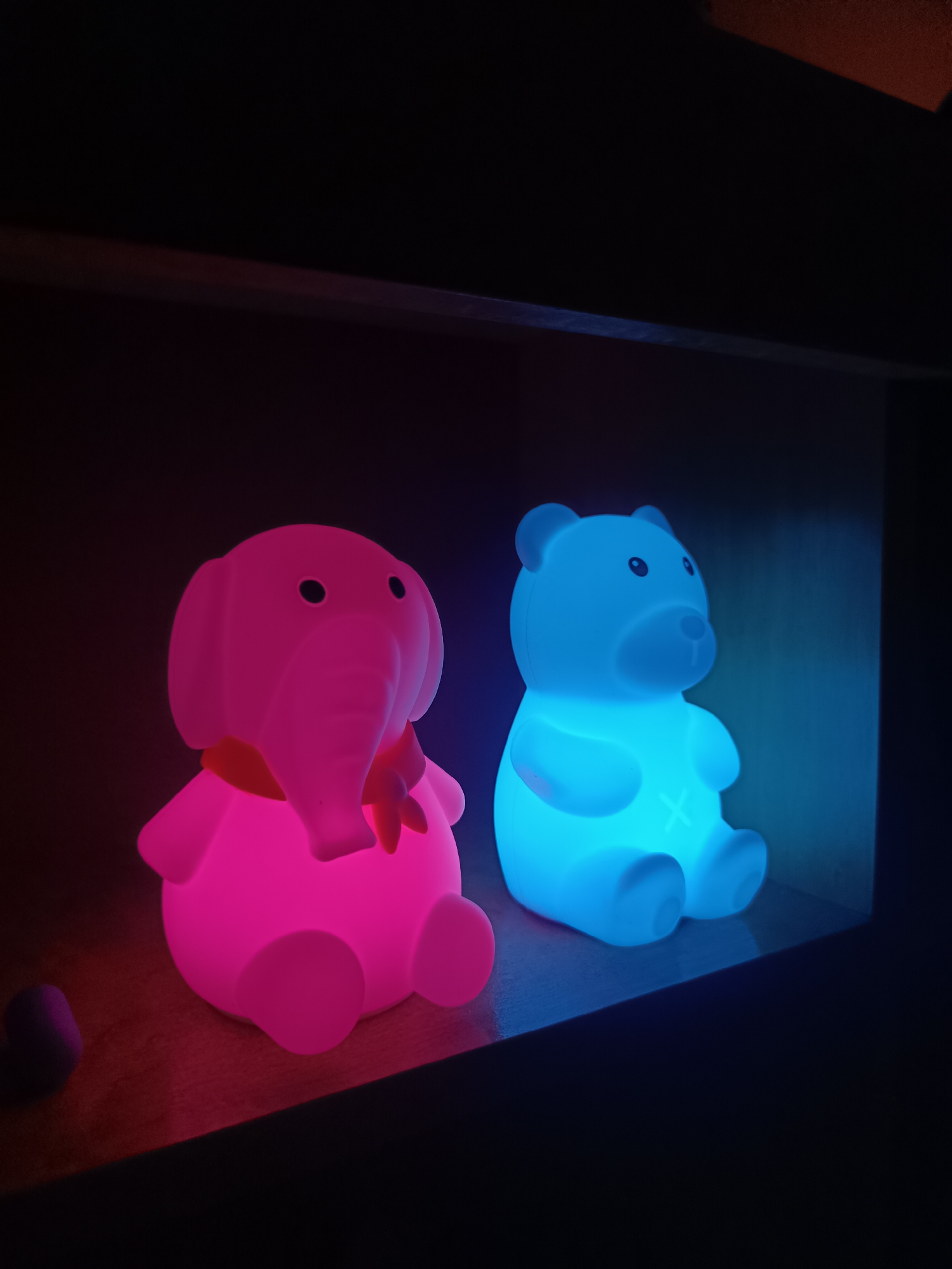 Two illuminated, cartoon-like figurines sit side-by-side on a dark shelf within a recessed space. The figurine on the left is pink, resembling an elephant wearing a scarf. The one on the right is blue and shaped like a bear. The background is dark, suggesting low ambient light. A small dark object is visible next to the pink elephant. The image location in the city of Cherkovitsa, Bulgaria, cannot be determined from the visual information provided.