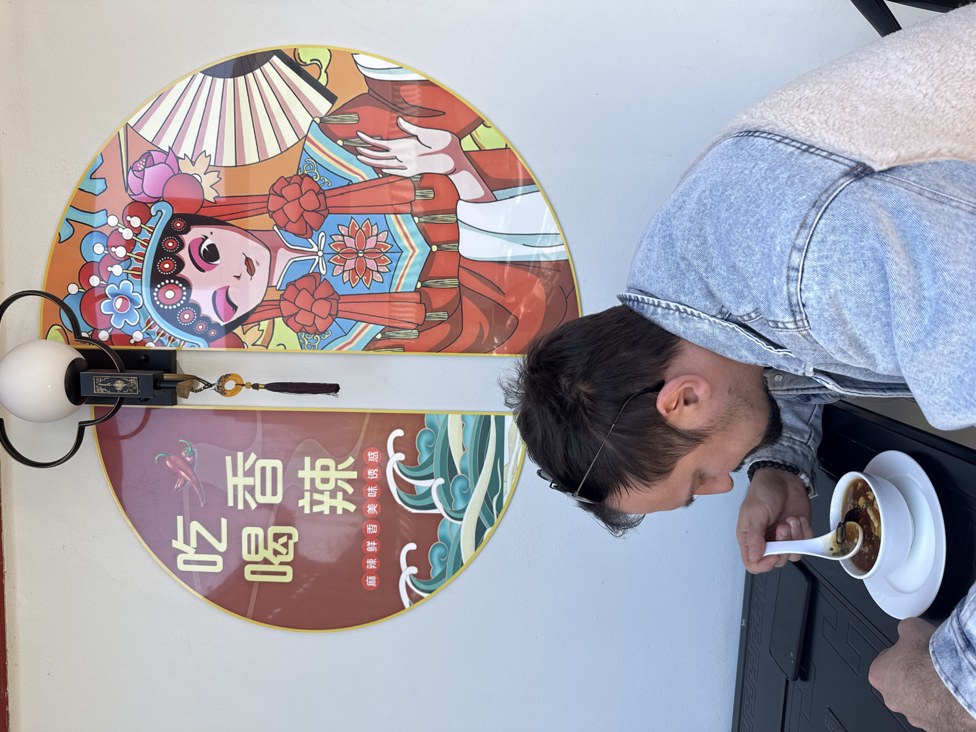 A man in a denim jacket with a sherpa collar is captured in a side profile, leaning over a dark table to eat soup from a white bowl. He uses a white spoon and has sunglasses pushed up onto his dark hair. The soup appears to be a dark, savory broth with various ingredients like mushrooms visible.
Behind him, on a light-colored wall, are two decorative semicircular panels, suggesting an Asian-themed eatery. The upper panel displays a vibrant, stylized illustration of a figure, likely a performer in traditional Chinese opera attire, adorned with an elaborate headdress and holding a fan. The artwork features a rich palette of red, orange, blue, pink, green, and yellow.
The lower panel, in a deep reddish-brown hue, includes Chinese characters and an illustration of a red chili pepper. The prominent characters read "吃喝" (chīhē), meaning "eat and drink," and "香辣" (xiānglà), meaning "fragrant and spicy." Below these, smaller text states "麻辣鲜香 美味诱惑" (málà xiānxiāng měiwèi yòuhuò), translating to "numbingly spicy, fresh and fragrant, delicious temptation." A decorative black light fixture with an amber-colored tassel hangs between the two wall art pieces.
The scene is brightly lit, indicating daytime. The man is engrossed in his meal, and the distinct decor points to a Chinese restaurant setting, located in Hainburg an der Donau, Austria.