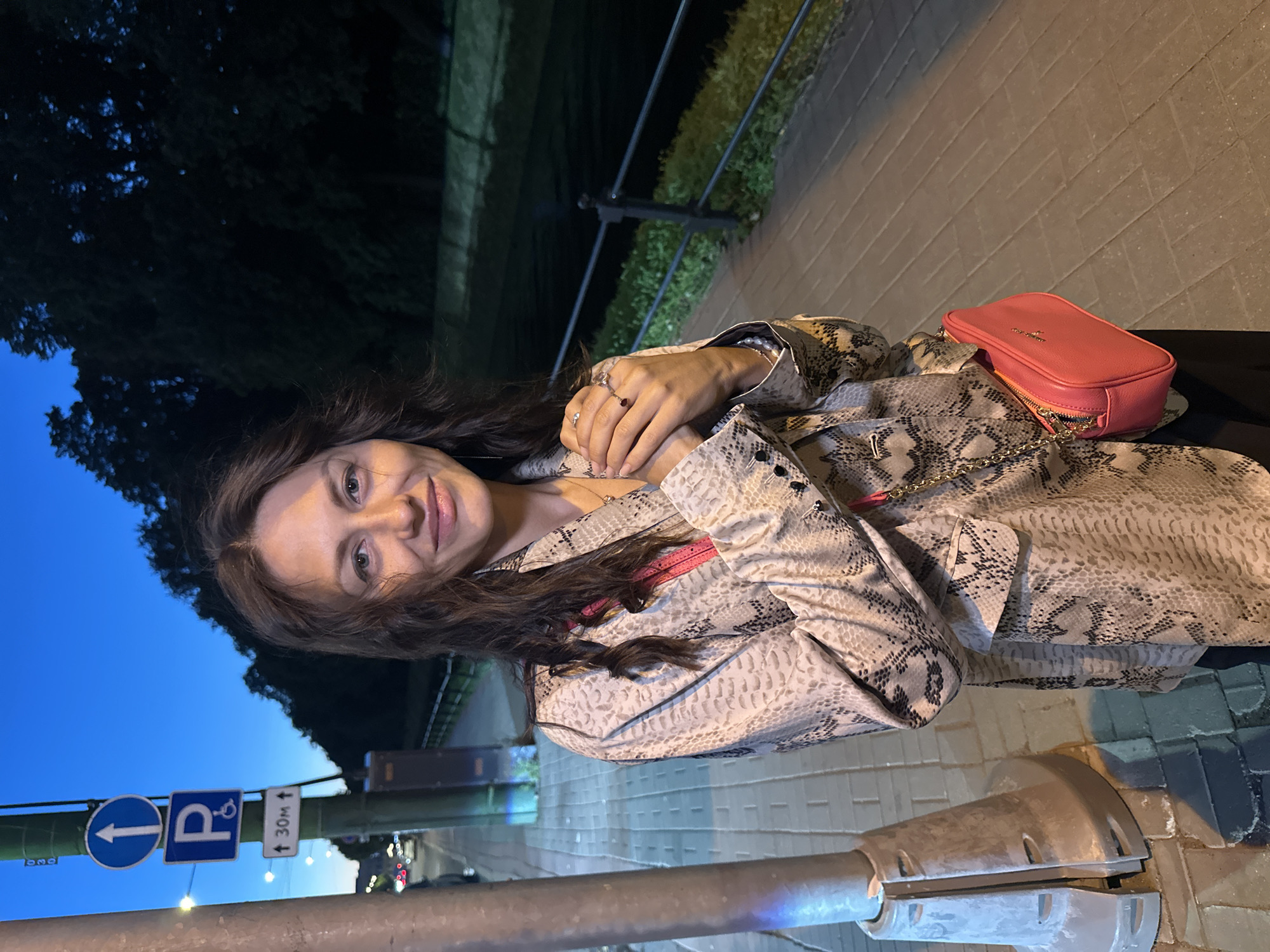A young woman with long, wavy brown hair and fair skin is standing on a brick sidewalk at dusk. She is smiling and holding her hands together in front of her chest. She is wearing a patterned jacket over a dark top and carrying a pink crossbody bag. To her left, a lamppost and several street signs are visible. The background features a body of water, trees, and a dark sky, suggesting an urban park or waterfront area in Saint Petersburg, Russia. The overall lighting indicates it is nighttime.
