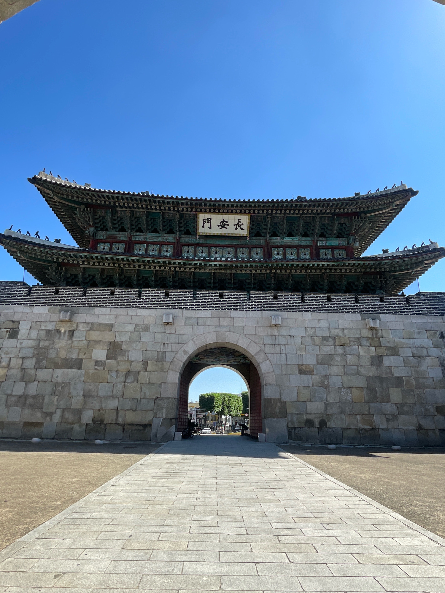 The image captures a frontal view of the Paldalmun Gate, the South Gate of Hwaseong Fortress in Suwon-si, South Korea. The gate is a two-story structure with a traditional Korean tiled roof, adorned with intricate wooden carvings and colorful paint. The roof features multiple eaves that curve upwards, typical of historical Korean architecture. Below the ornate upper structure is a massive stone wall, constructed from large, irregular blocks. An arched gateway pierces through this stone wall, leading into the fortress grounds. The pathway leading to the arch is paved with rectangular stones. Beyond the arch, a glimpse of greenery and some people can be seen, suggesting an active tourist area. The sky above is a clear, bright blue. The overall scene is bathed in bright sunlight, casting shadows on the ground.