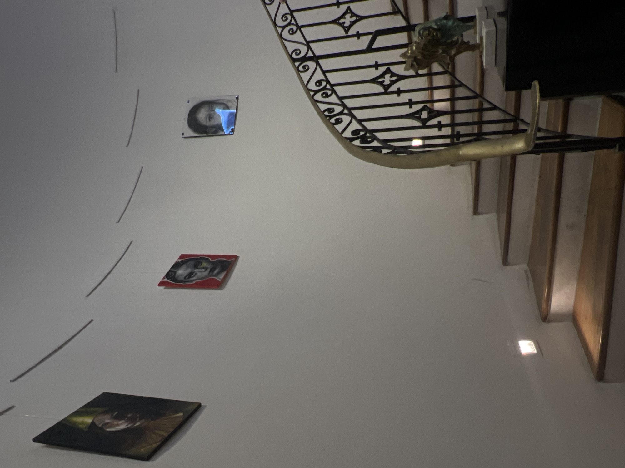 The image shows an upward view of a staircase and a section of an adjacent wall adorned with artworks. The staircase features ornate, black metal railings with gold accents, ascending in a curve. The wall to the left is painted a neutral color and displays three framed portraits suspended at different heights. The subjects in the portraits appear to be faces. The scene is indoors and illuminated with soft lighting. While it is unclear from this image alone that this is Mexico City, Mexico.