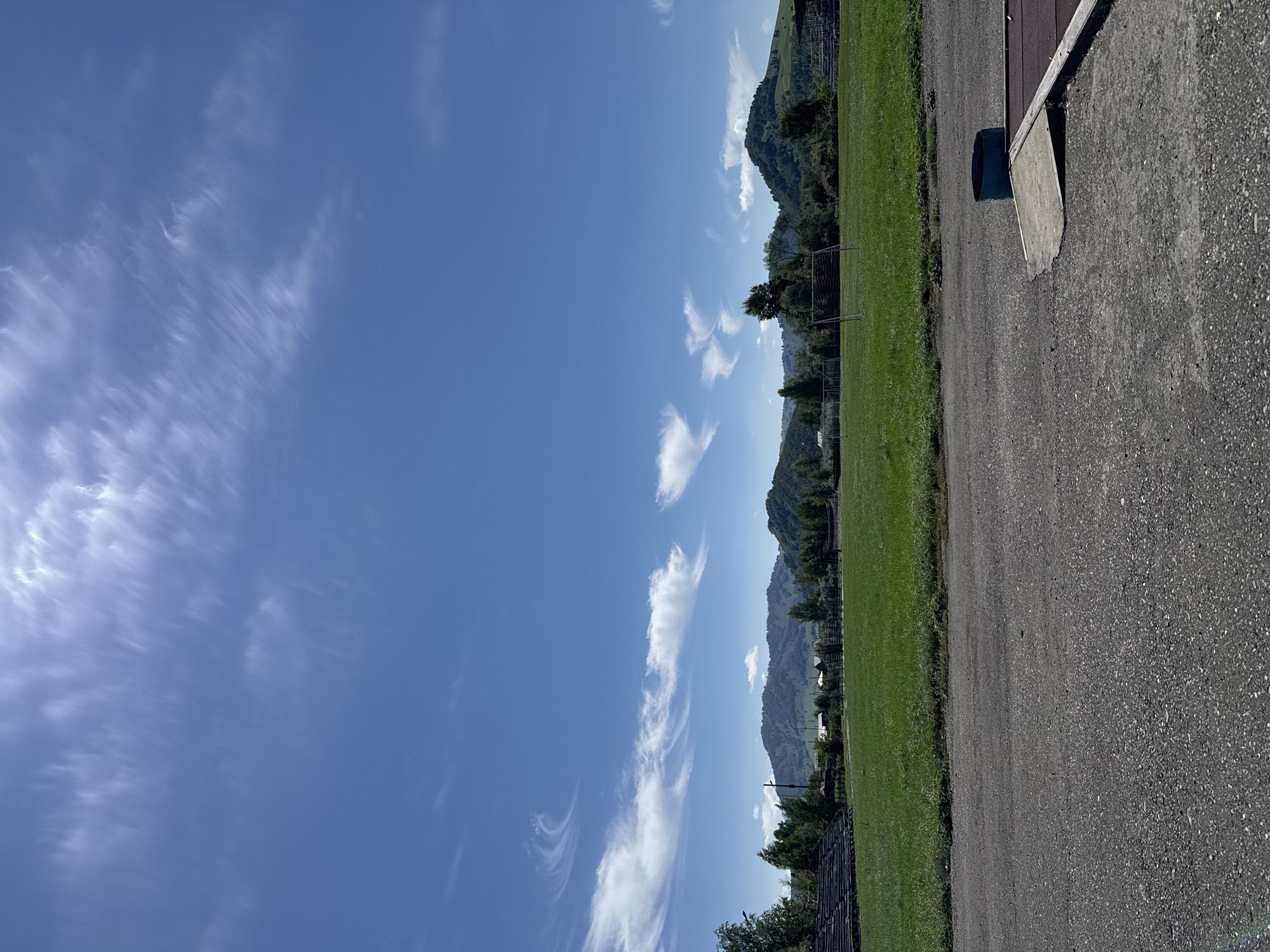 The image captures a wide view of a green sports field, likely a soccer field given the presence of two goals. Beyond the field, rolling hills covered in lush green vegetation and trees form a natural backdrop. The sky above is a vibrant blue, sparsely dotted with wispy white clouds, suggesting fair weather. The foreground of the image consists of a paved surface, possibly a track or pathway, with a dark blue cylindrical object and a concrete structure visible. To the left of the field, wooden bleachers are partially visible, indicating a spectator area. Scattered houses and fences are also present in the middle ground, suggesting a rural or semi-rural setting. The scene appears to be in the village of Soloneshnoye, Russia, known for its scenic natural beauty.
