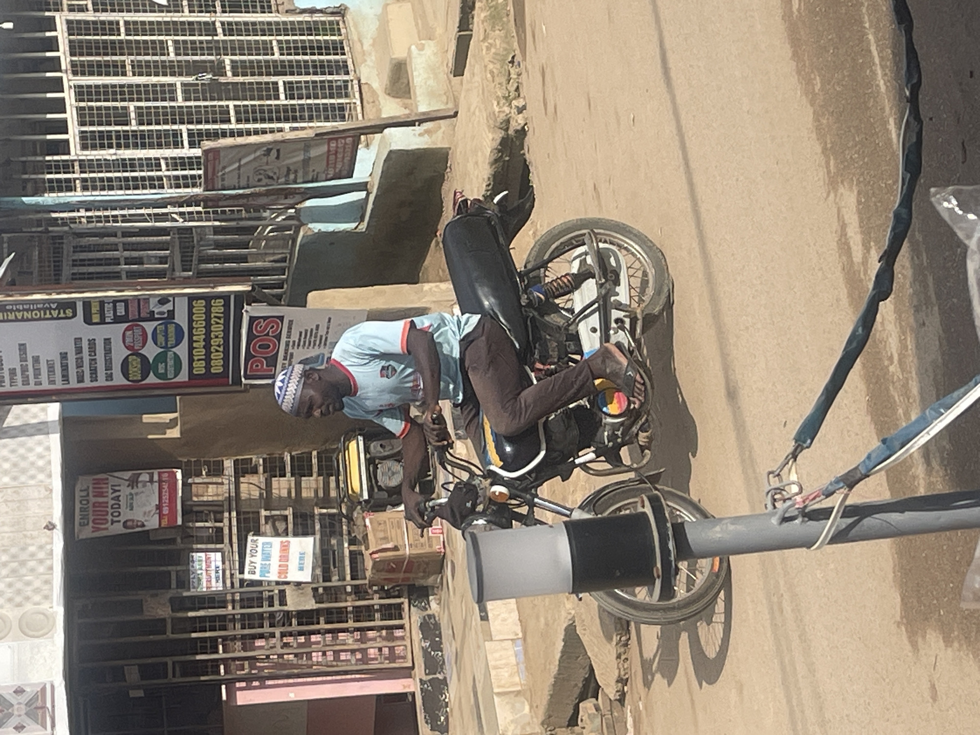 A man rides a motorcycle down a street in Abuja, Nigeria. The man is wearing a light blue shirt with an orange collar and a white and blue patterned cap. He is also wearing brown pants and sandals. The motorcycle is black with yellow and blue accents. It has a large, cylindrical object attached to the front. In the background, there are buildings with metal bars on the windows and various signs, including one that says "ENROLL YOUR NIN TODAY!" and another that says "POS." The street appears to be paved with dirt and gravel, and there is a lamppost with wires attached to it in the foreground. The overall impression is that of a busy street scene in an urban environment.
